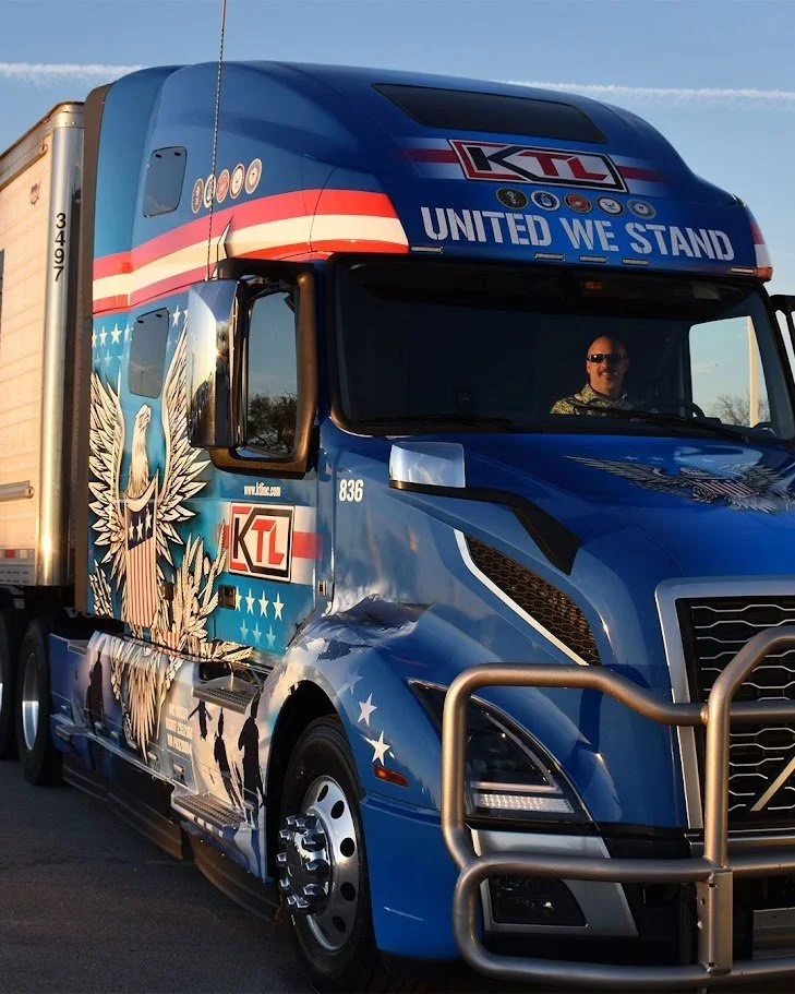 Blue semi-truck with patriotic American eagle mural and the slogan 'United We Stand' on the front, driving on a road during daytime.