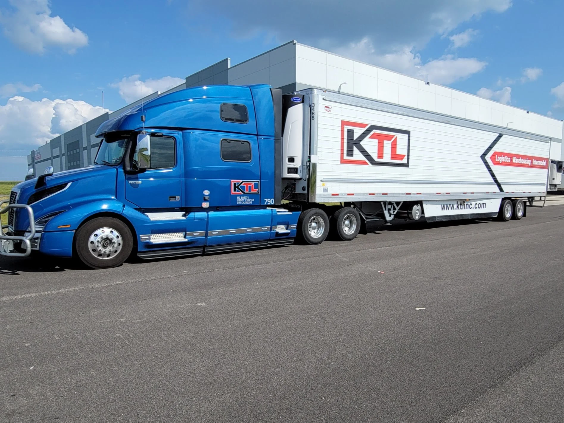 Blue semi-truck with a white trailer parked on a paved road under a partly cloudy sky near a modern building.