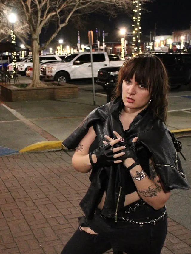 A woman outside at night, holding the leather jacket wrapped around her shoulders.