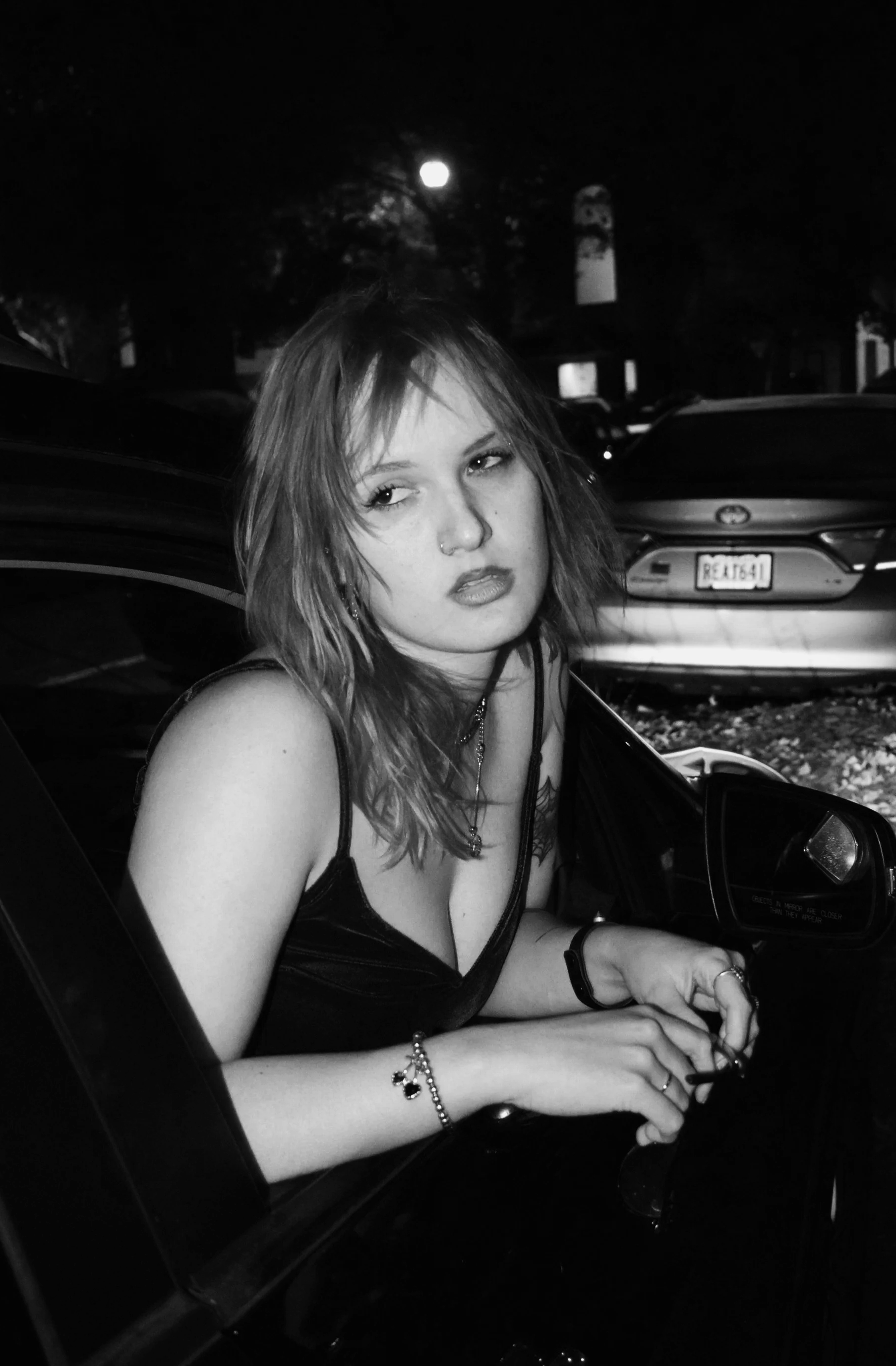 A young woman with shoulder-length hair and a nose piercing sitting in a black car at night, with parked cars and a full moon in the background.