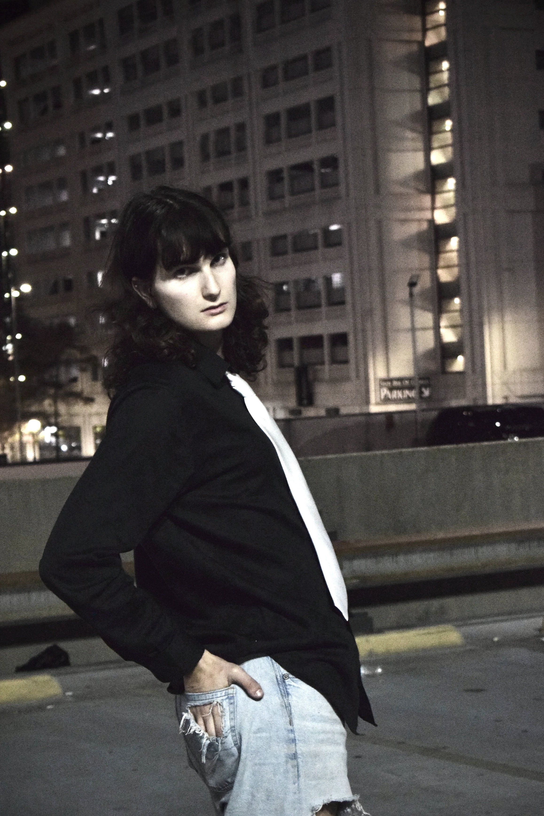 A woman with dark curly hair wearing a black jacket and ripped jeans standing in an urban parking lot at night, with tall buildings and city lights in the background.