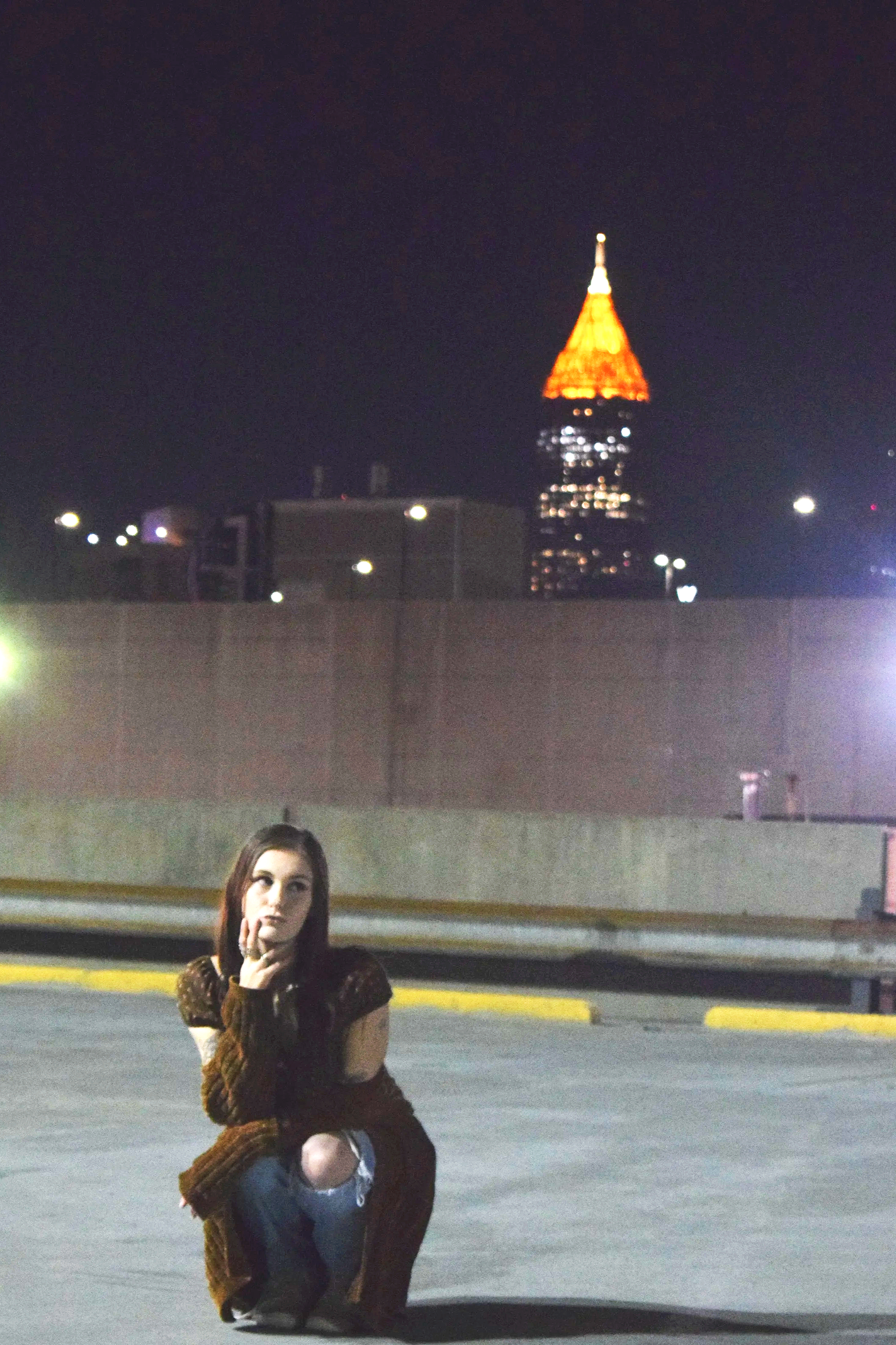 A woman crouching on a rooftop at night with the city skyline and a lit tower in the background.
