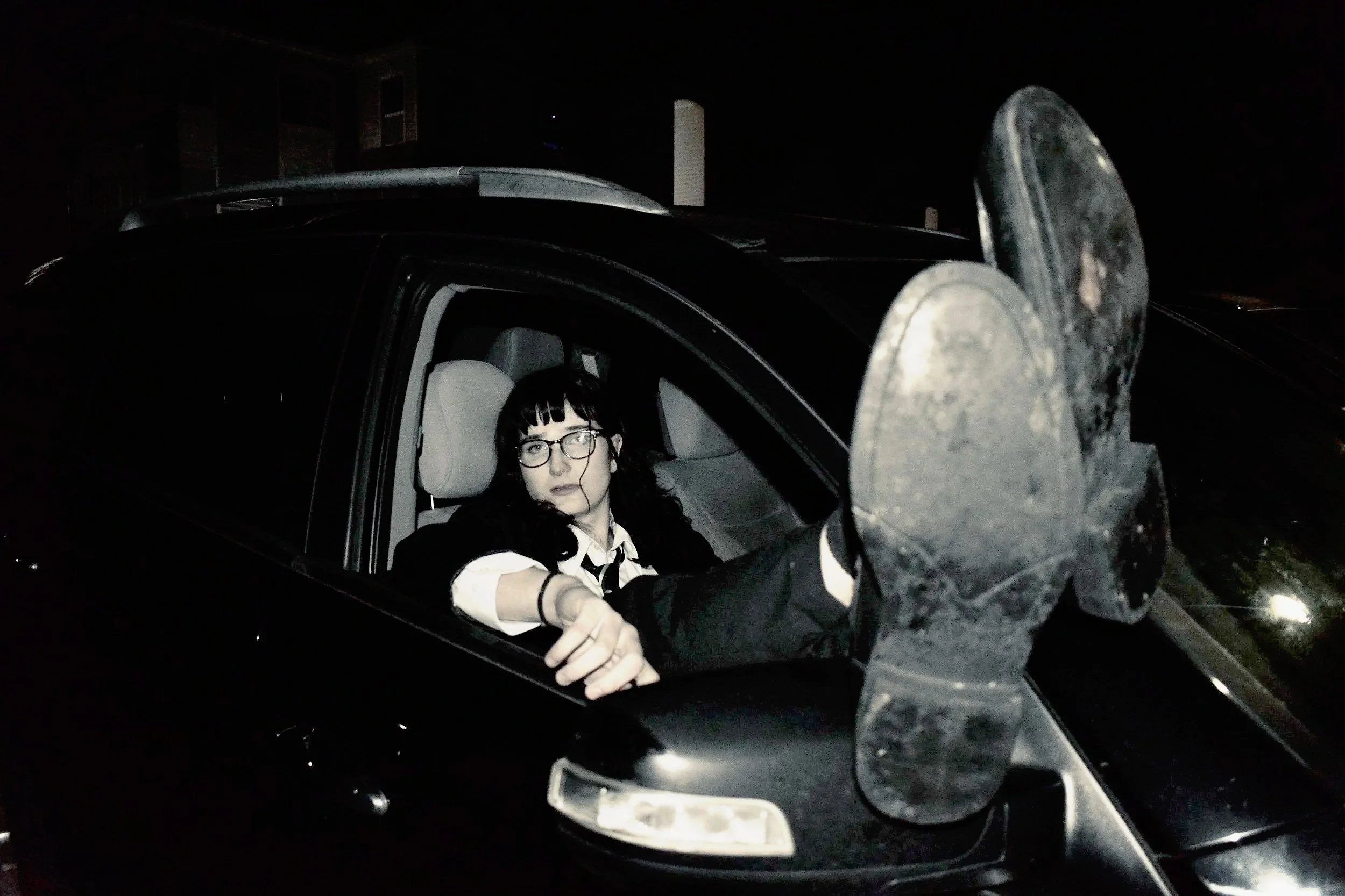 A woman with glasses and dark hair sits in the driver's seat of a car at night, with her foot extended out of the window, showing the sole of her shoe.