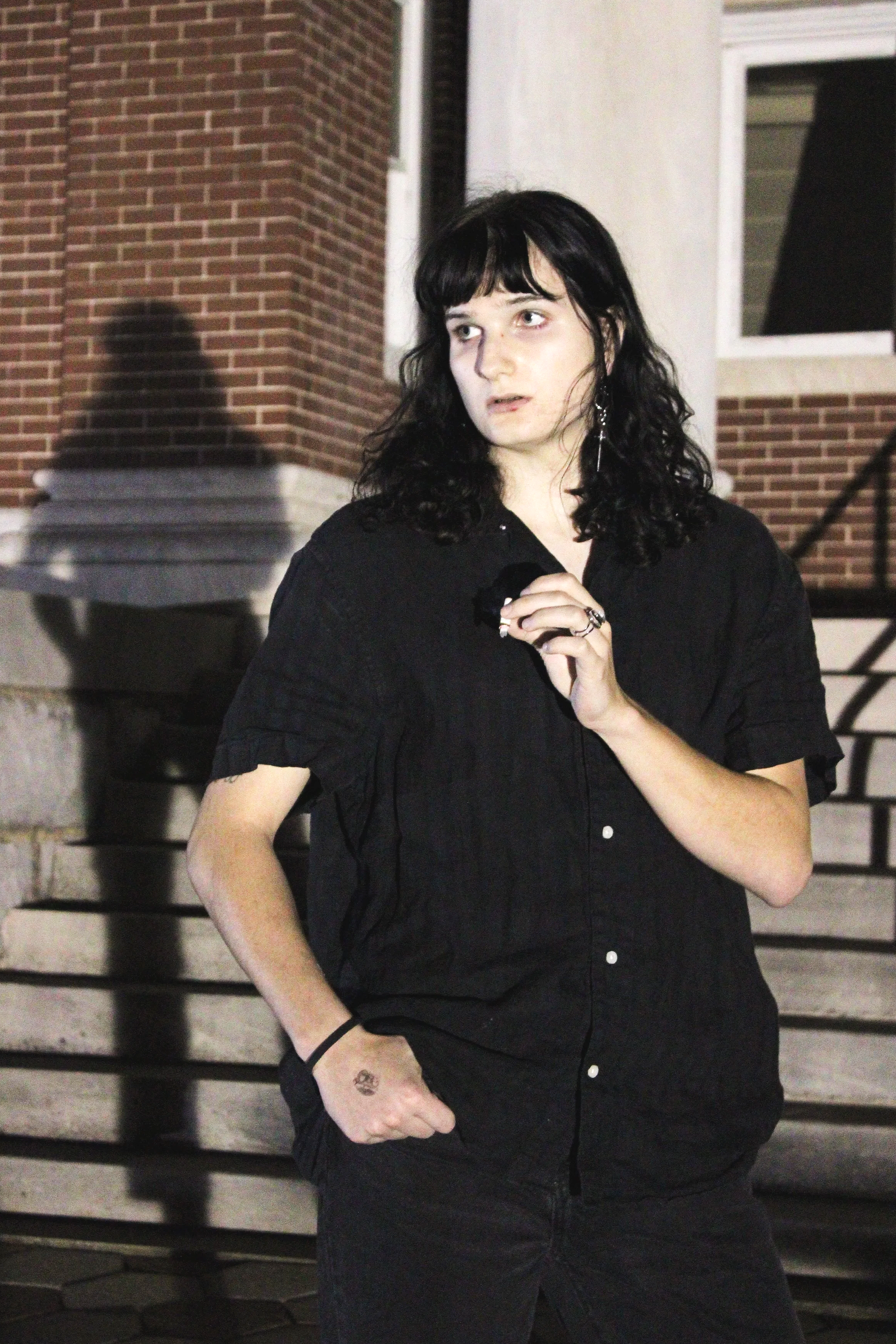 A young woman with black wavy hair, wearing a black short-sleeved shirt, standing outside near brick stairs at night with a shadow cast on the wall behind her.