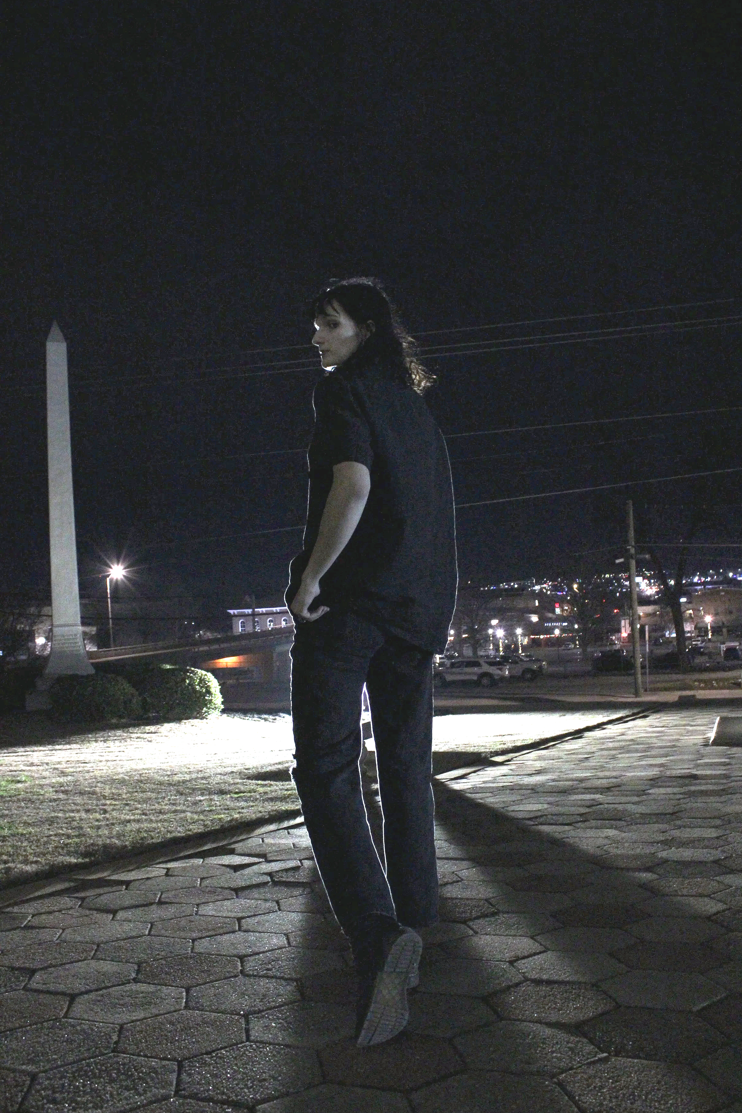 A woman dressed in black pants and a black jacket standing on a brick sidewalk at night, with City skyline and streetlights in the background.