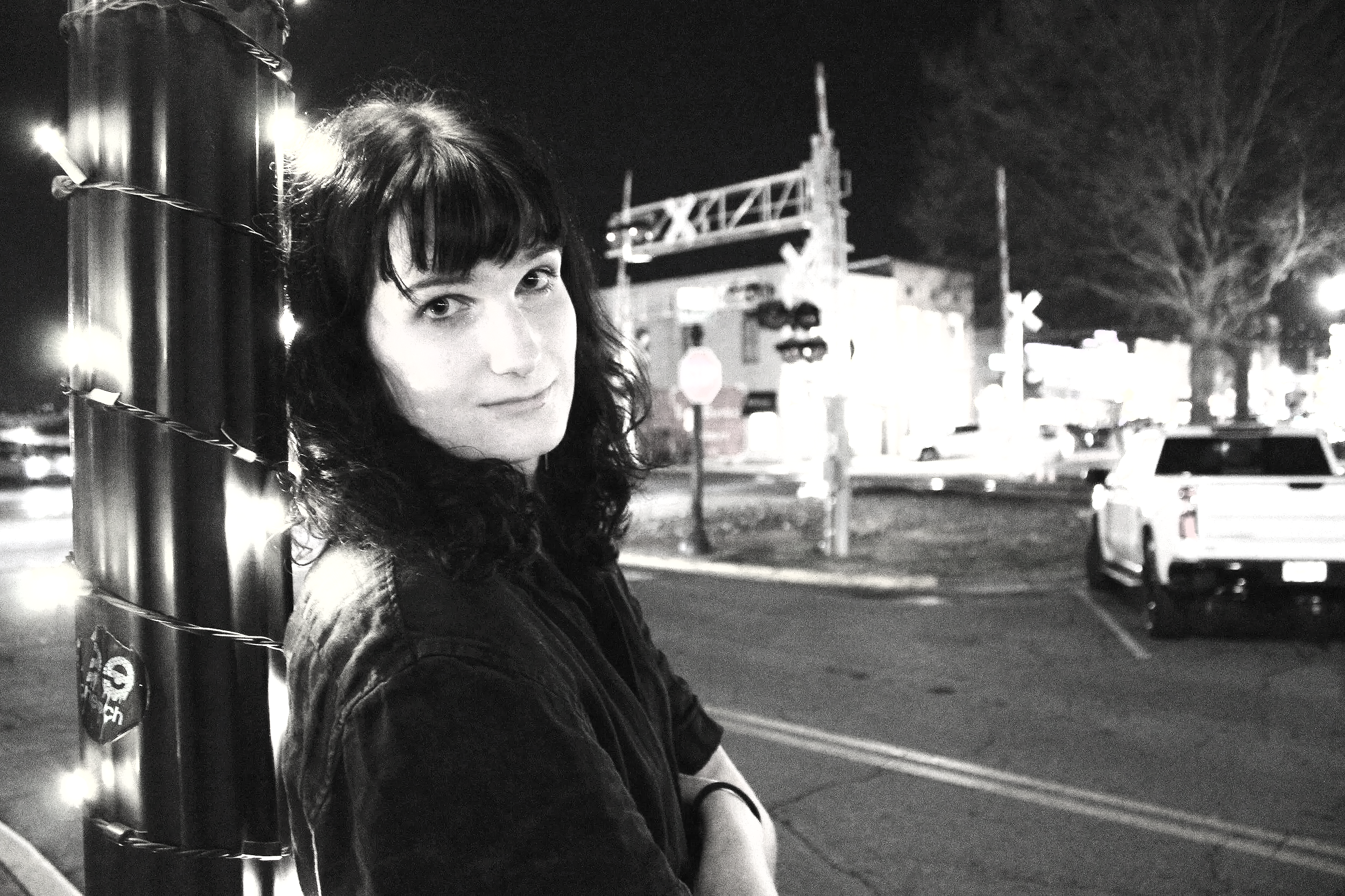 A woman with dark curly hair and bangs stands on a city street at night, leaning against a pole decorated with string lights. The background features blurred cars, buildings, and a traffic signal.