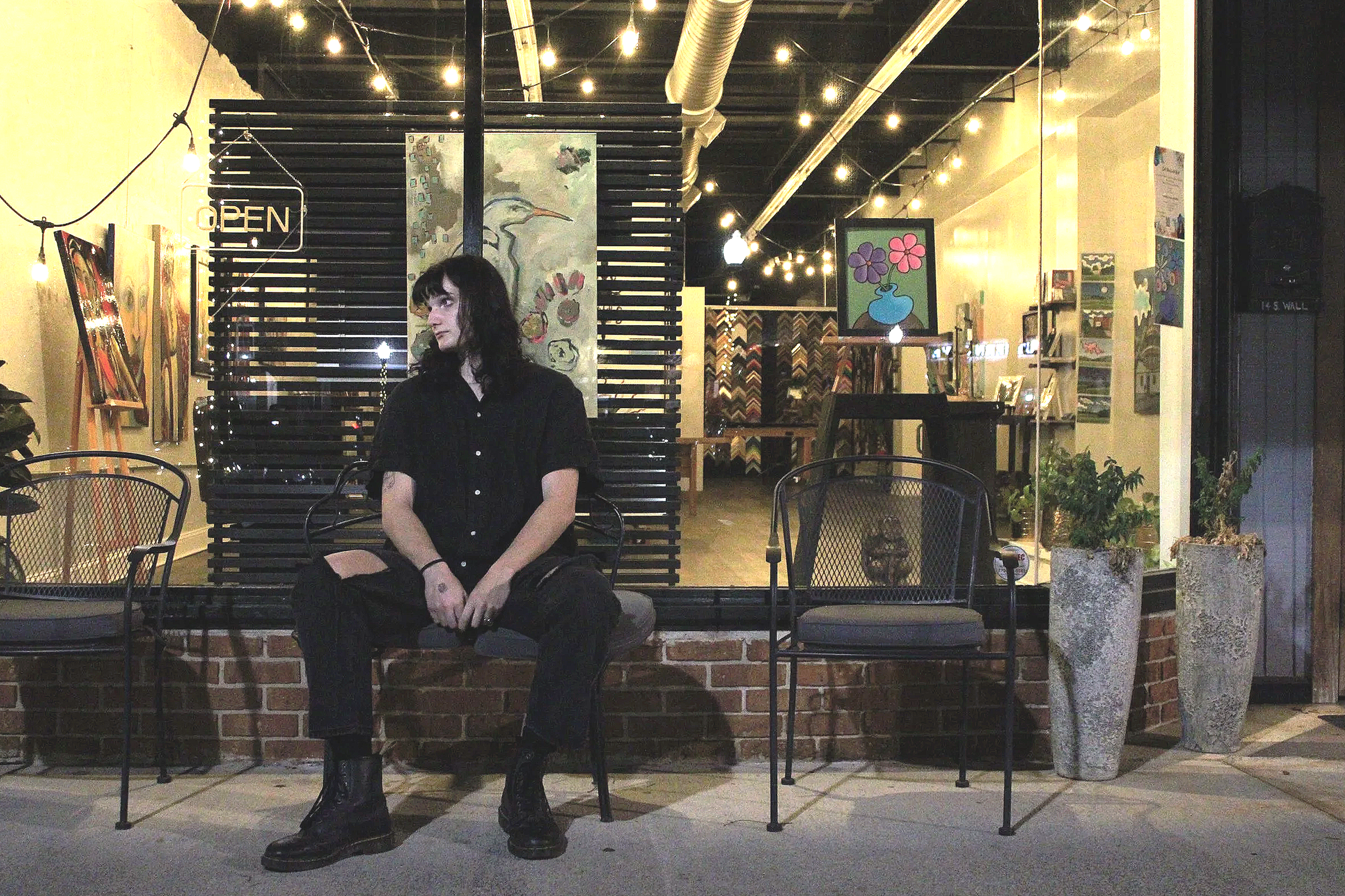 A woman with dark hair sitting alone on a bench outside an art gallery or cafe at night. The gallery has art on the walls, potted plants, and string lights overhead.