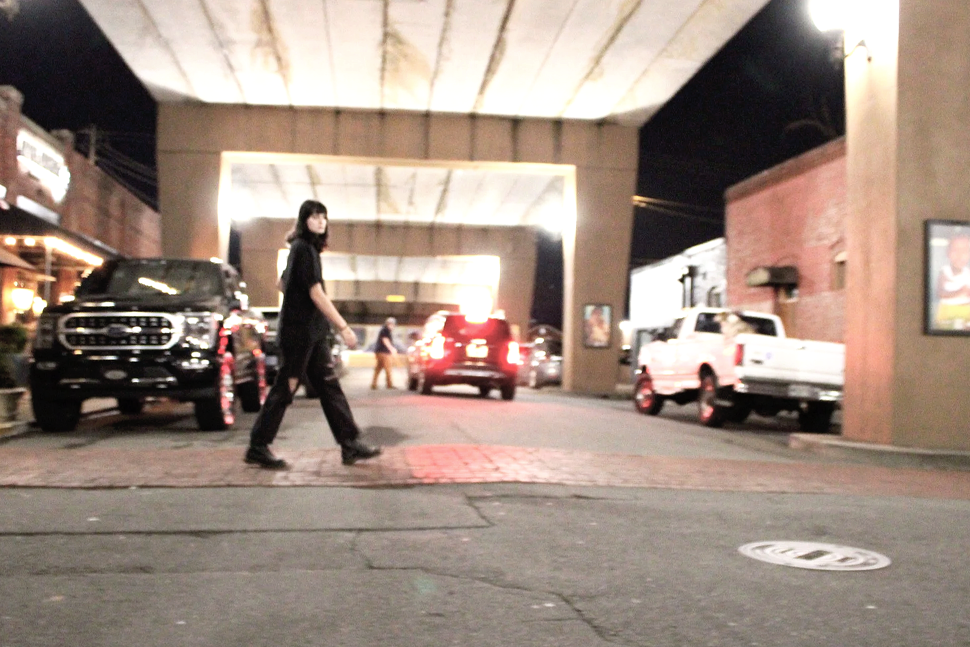 A woman in black walking on a city sidewalk at night, with cars parked and moving in the background under a large bridge or overpass.