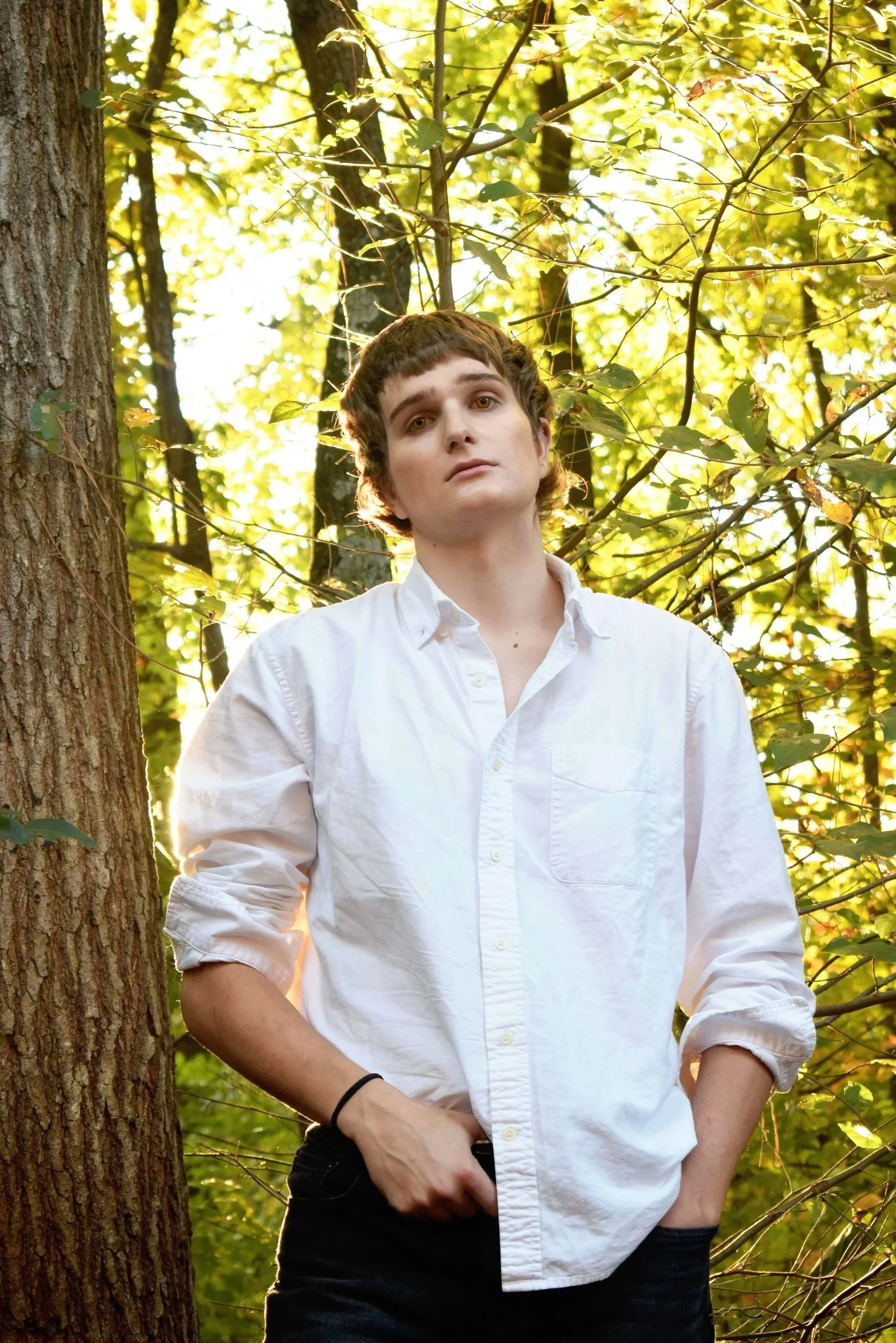 A young person standing outdoors in front of trees with green leaves, wearing a white button-up shirt with relaxed sleeves, black pants, and a black wristband, with one hand in the pocket and the other resting on their hip, looking at the camera.