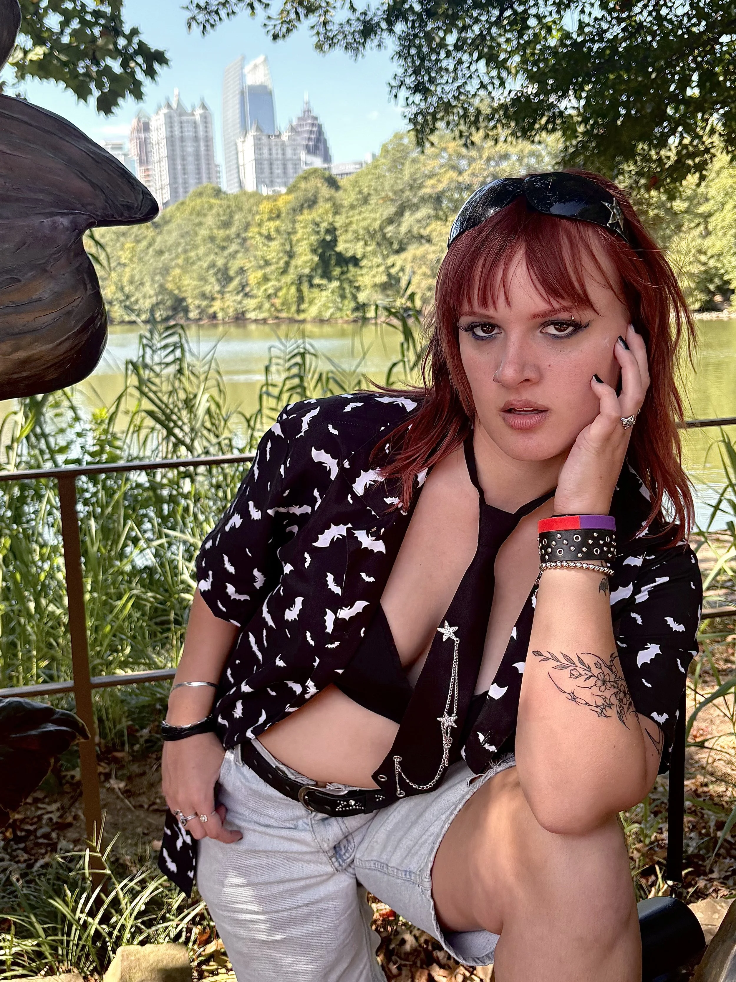 A woman with red hair, tattoos, and wearing sunglasses on her head is sitting outdoors near a lake with trees and a city skyline in the background. She is wearing a black and white patterned shirt and shorts, with jewelry and wristbands.