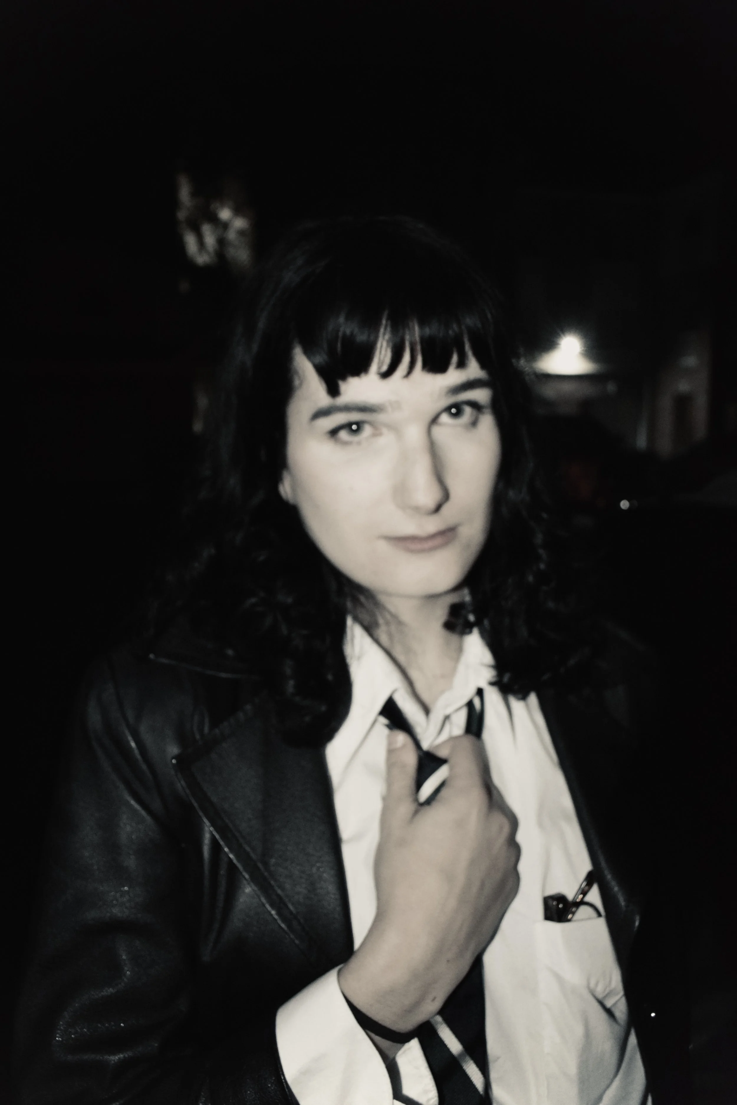 A woman with shoulder-length dark hair, wearing a white shirt, black tie, and black leather jacket, standing outdoors at night.