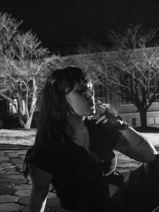 A woman sitting outside smoking at night in black-and-white.