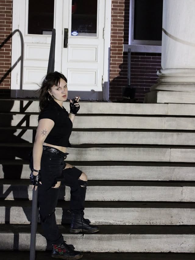 A woman smokes a cigarette while perching on stairs outside at night.