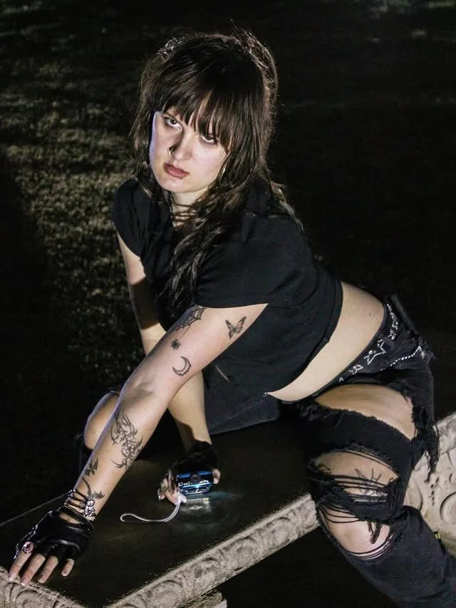 A woman on a bench outside at night, staring up at the camera.