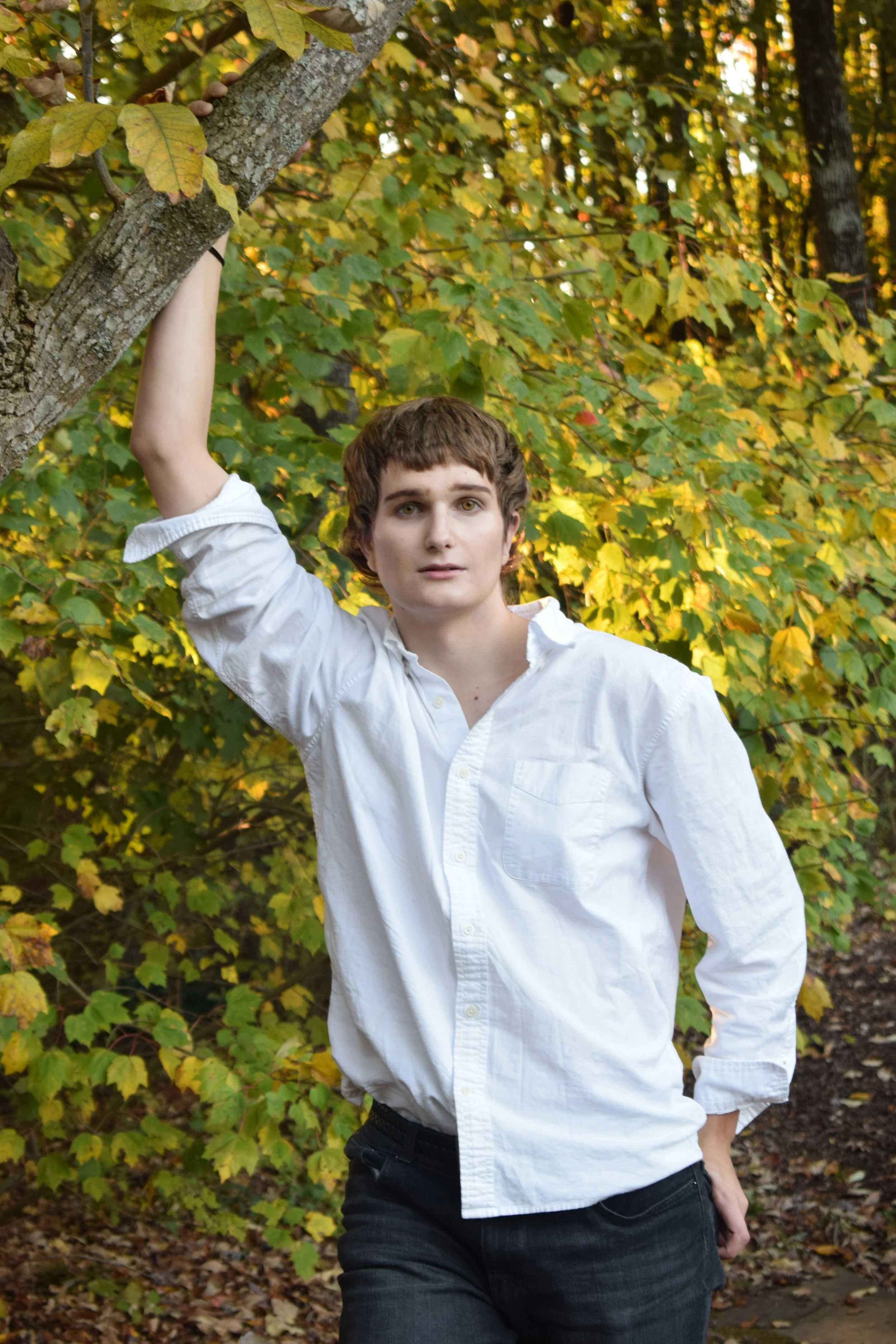 A young person with short brown hair in a white button-up shirt, standing outdoors in an autumn forest, holding onto a tree branch with one hand.