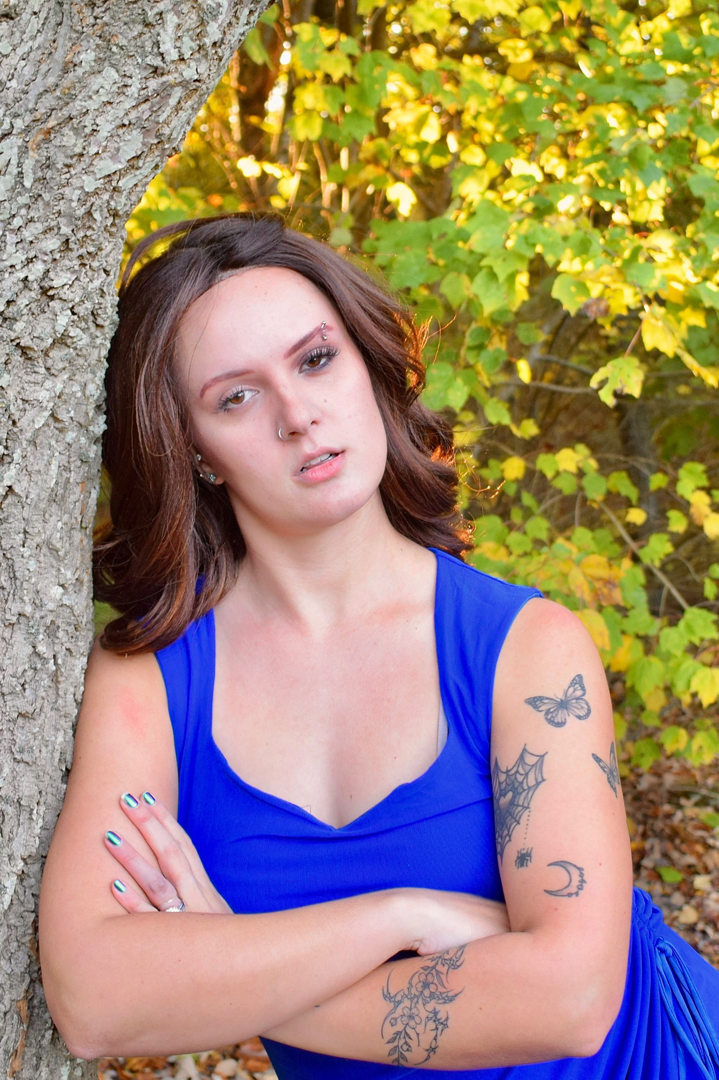 Young woman with brown hair and tattoos on her arms, wearing a blue sleeveless dress, leaning against a tree in a forest with yellow and green leaves.