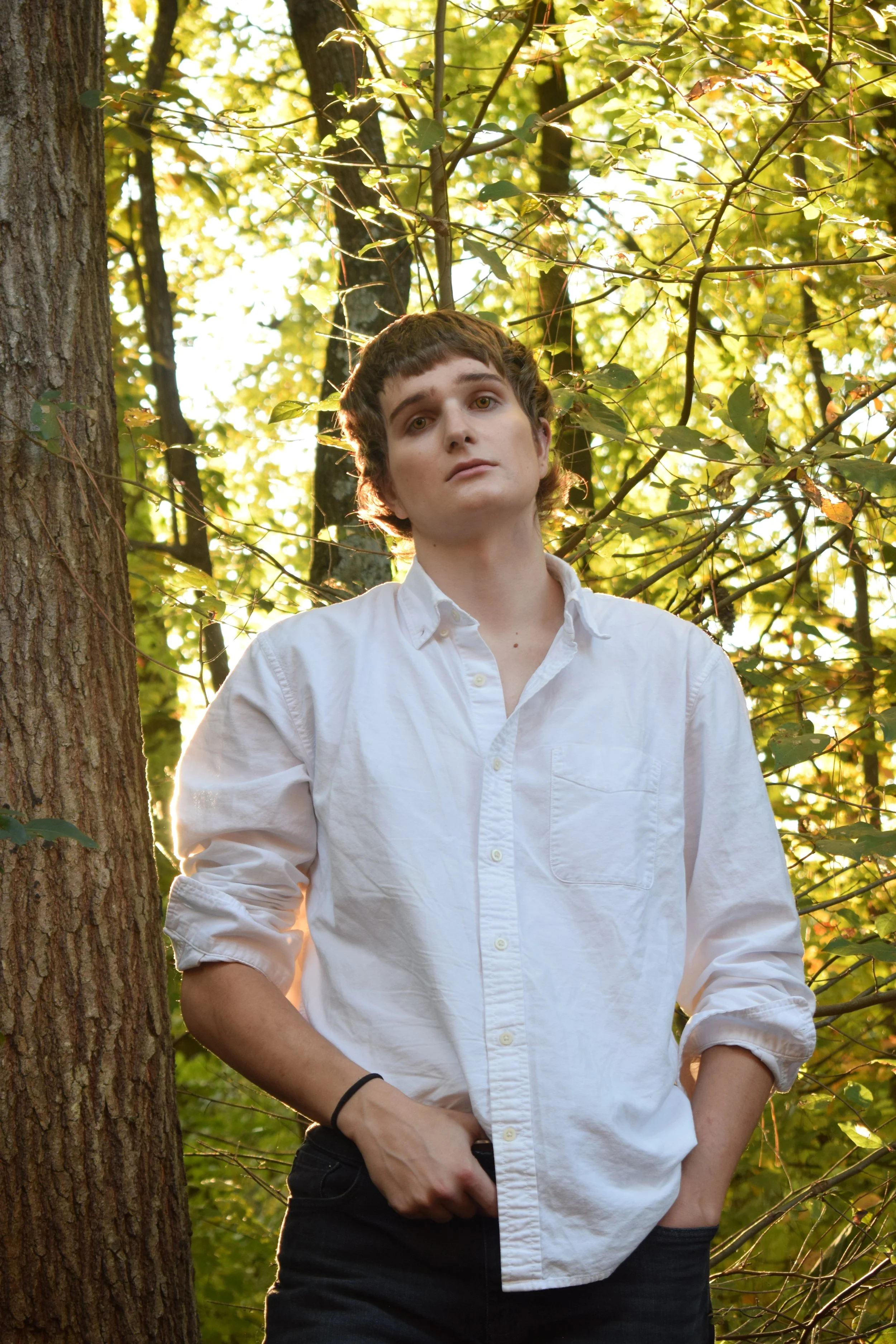 A young person standing outdoors in front of trees with green leaves, wearing a white button-up shirt with relaxed sleeves, black pants, and a black wristband, with one hand in the pocket and the other resting on their hip, looking at the camera.