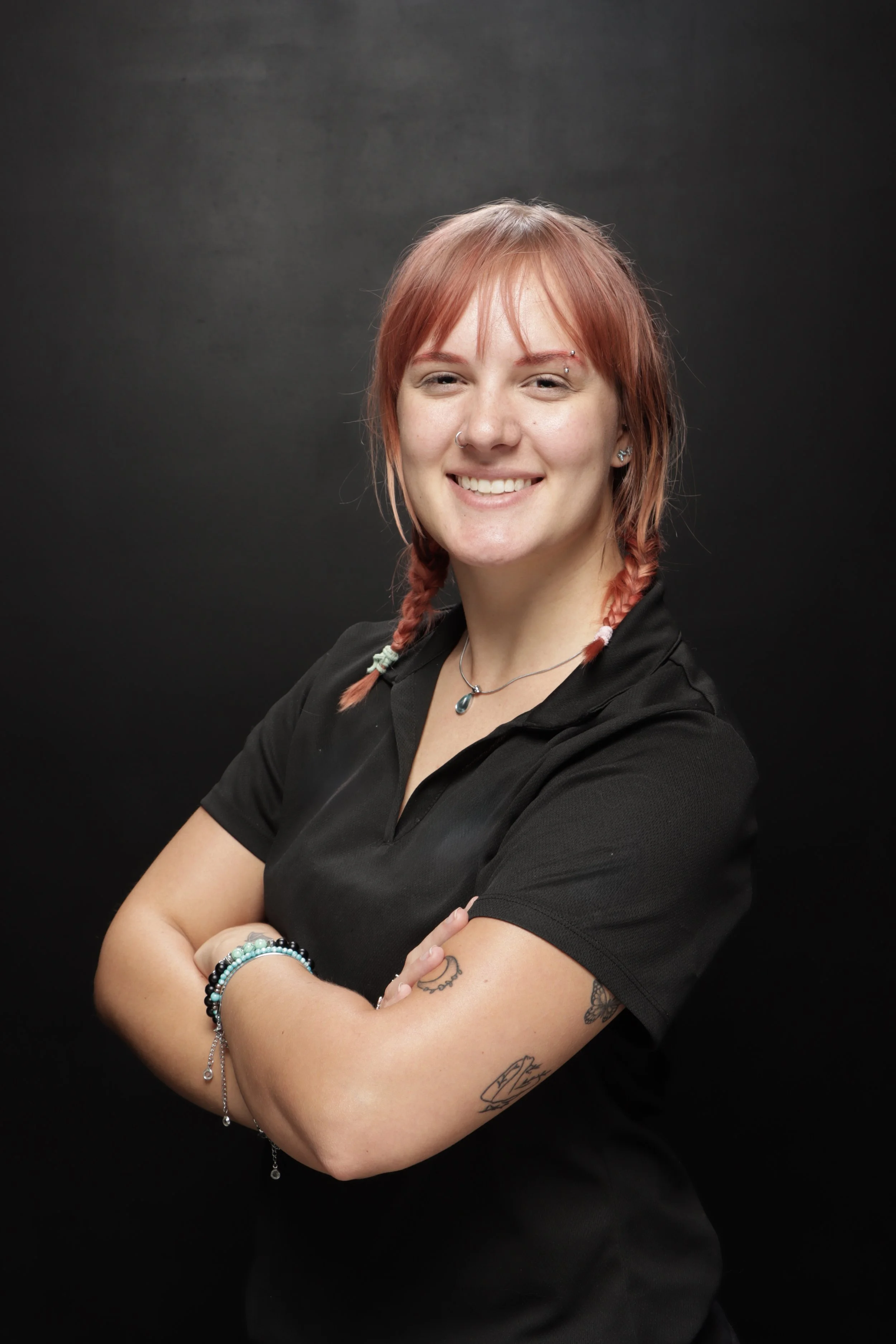 A smiling woman with light skin, pink hair in braids, wearing a black polo shirt, jewelry, and tattoos, standing with crossed arms against a dark background.