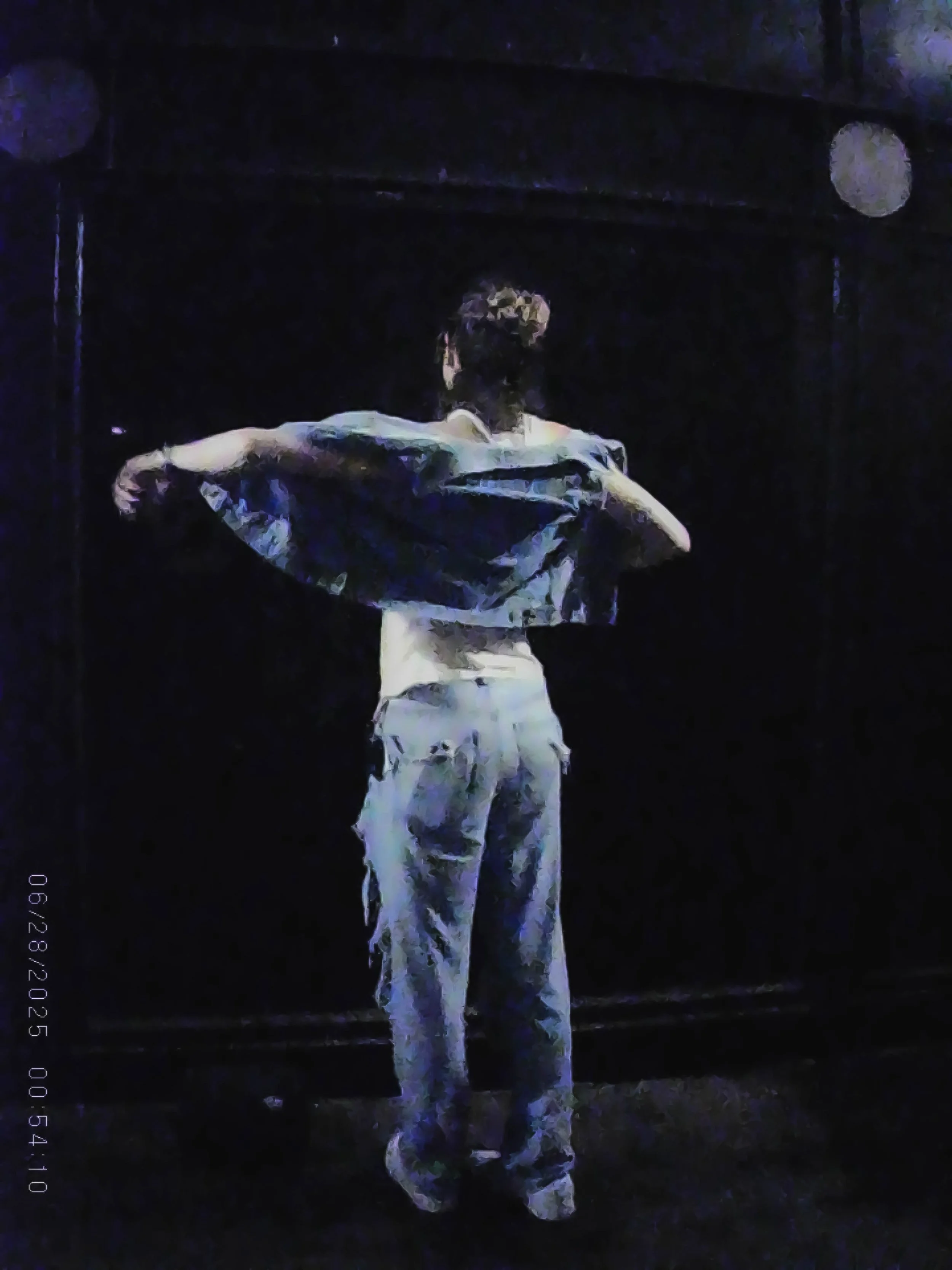 A person with short hair in loose clothing, pulling a jacket or shirt over their head, standing against a dark background.