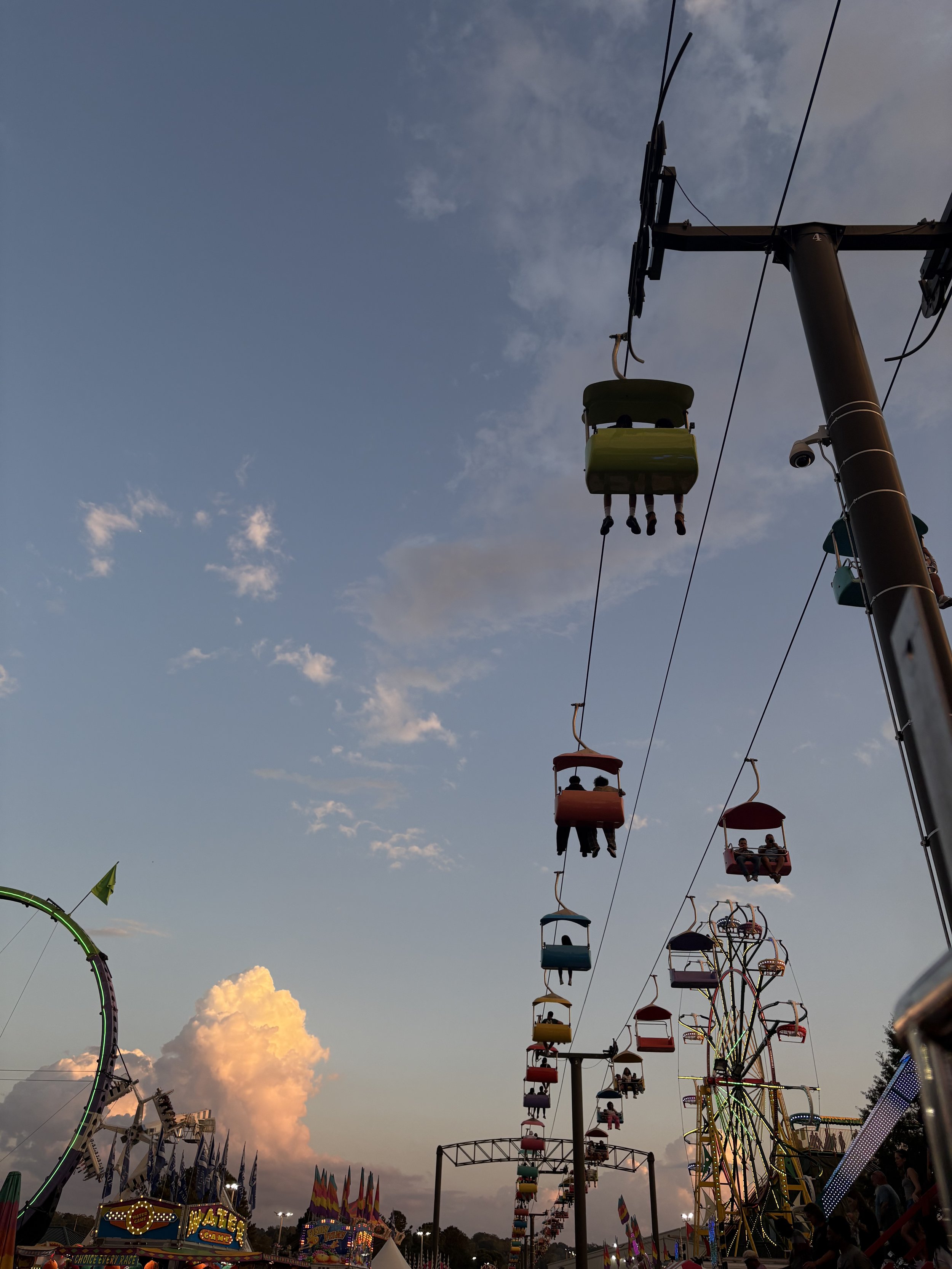 Ferris wheel and amusement park rides at sunset during a fair, with colorful gondolas, a carnival game, and a large cloud in the sky.
