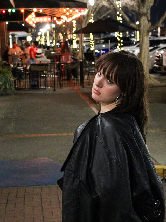A woman holds her leather jacket over her shoulder outside at night with a crowded restaurant in the background.