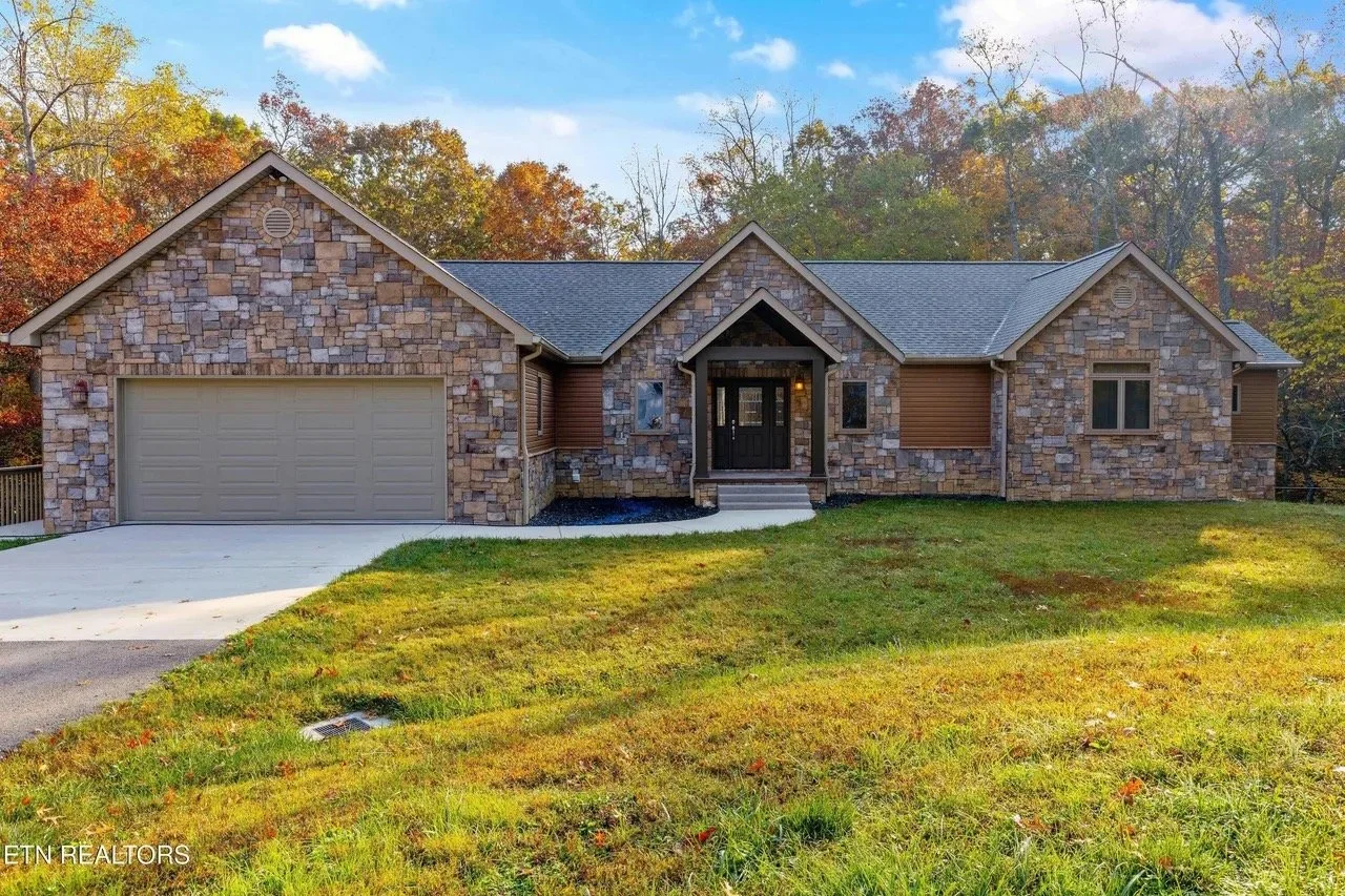 A house with a stone exterior, a front porch, a two-car garage, and a sloped lawn in front. The house is surrounded by trees with fall foliage.