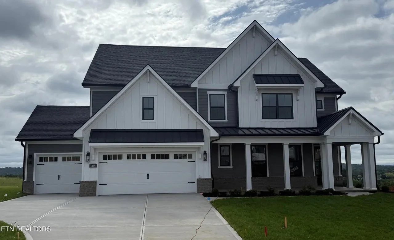 Front view of a modern, two-story house with a tripartite garage door, dark gray and white exterior, and a covered porch, set against a cloudy sky.