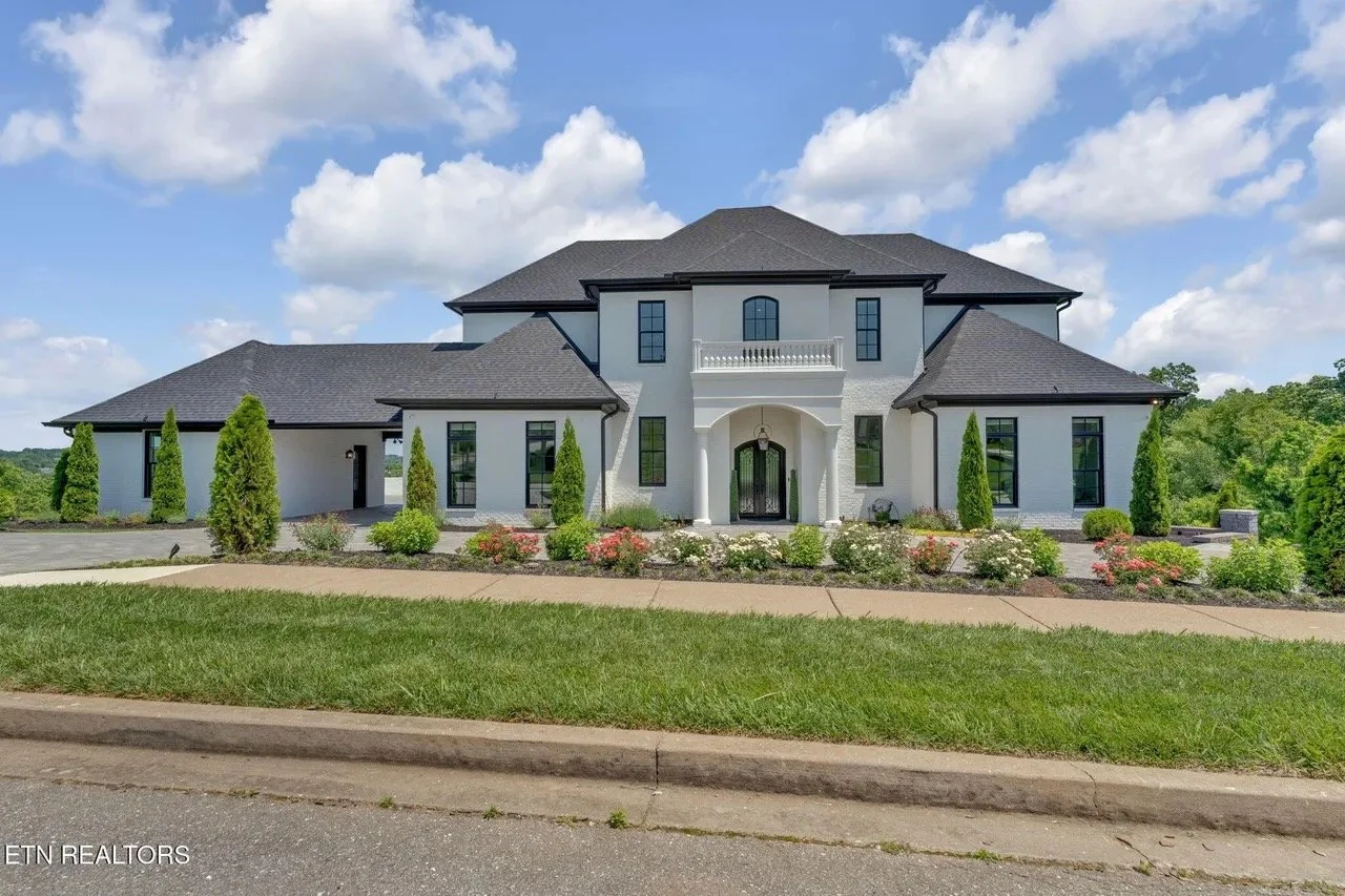 Large modern house with white walls and black roof, surrounded by landscaped garden with shrubs and trees, under a blue sky with clouds.