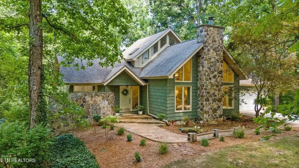 A quaint, green and stone house surrounded by trees and a landscaped yard with small plants, and a stone pathway leading to the front door.
