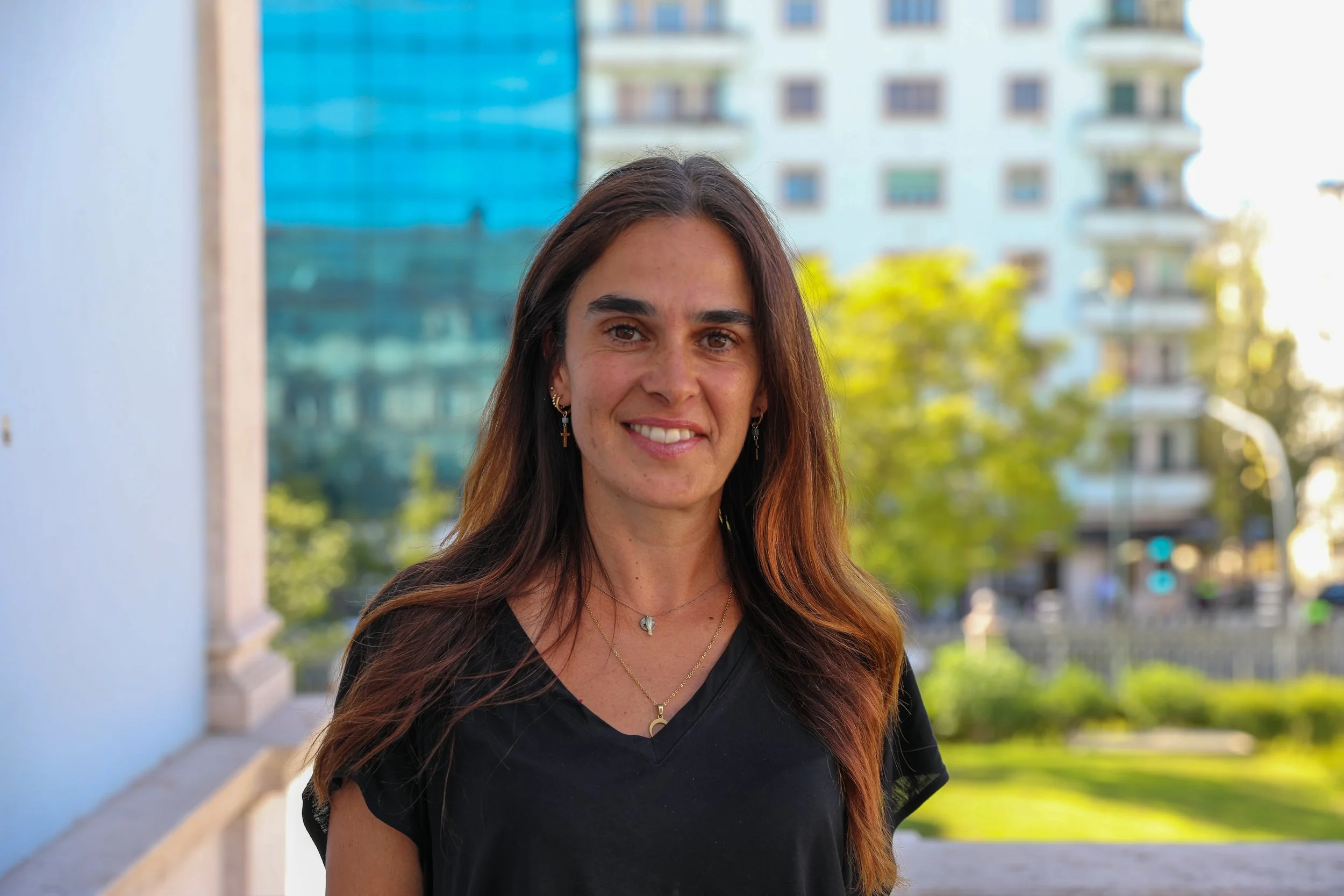 Smiling woman with long hair wearing a black shirt and standing outdoors with buildings and greenery blurred in the background.