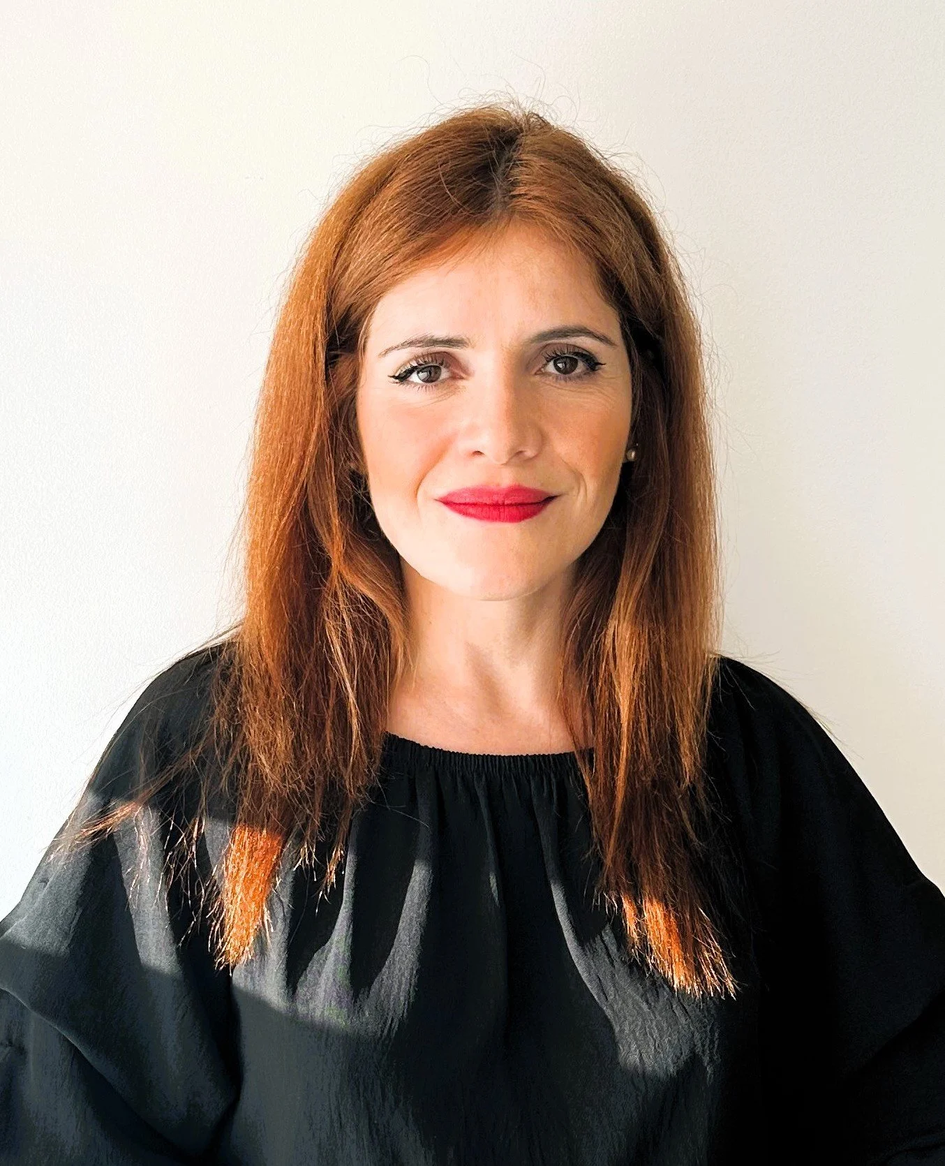 Portrait of a woman with long red hair and a black top, smiling, against a plain white background.