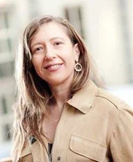 Person smiling with long hair, wearing earrings and a light-colored jacket.