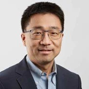 Man wearing a suit and glasses with a neutral expression against a plain background.