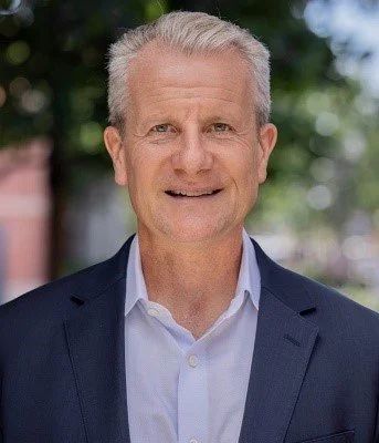 A middle-aged man in a suit standing outdoors with a blurred background of trees and buildings.