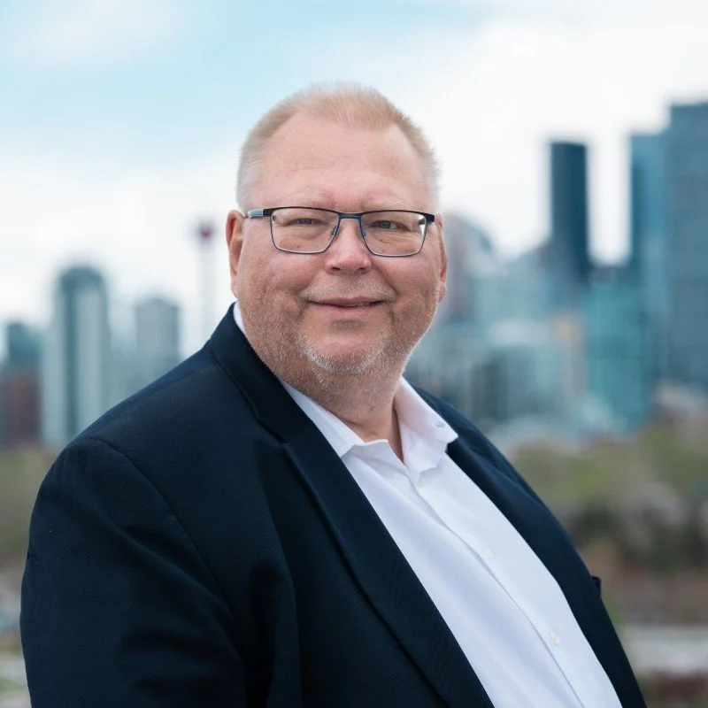 A man in a suit and glasses smiling with a cityscape in the background.