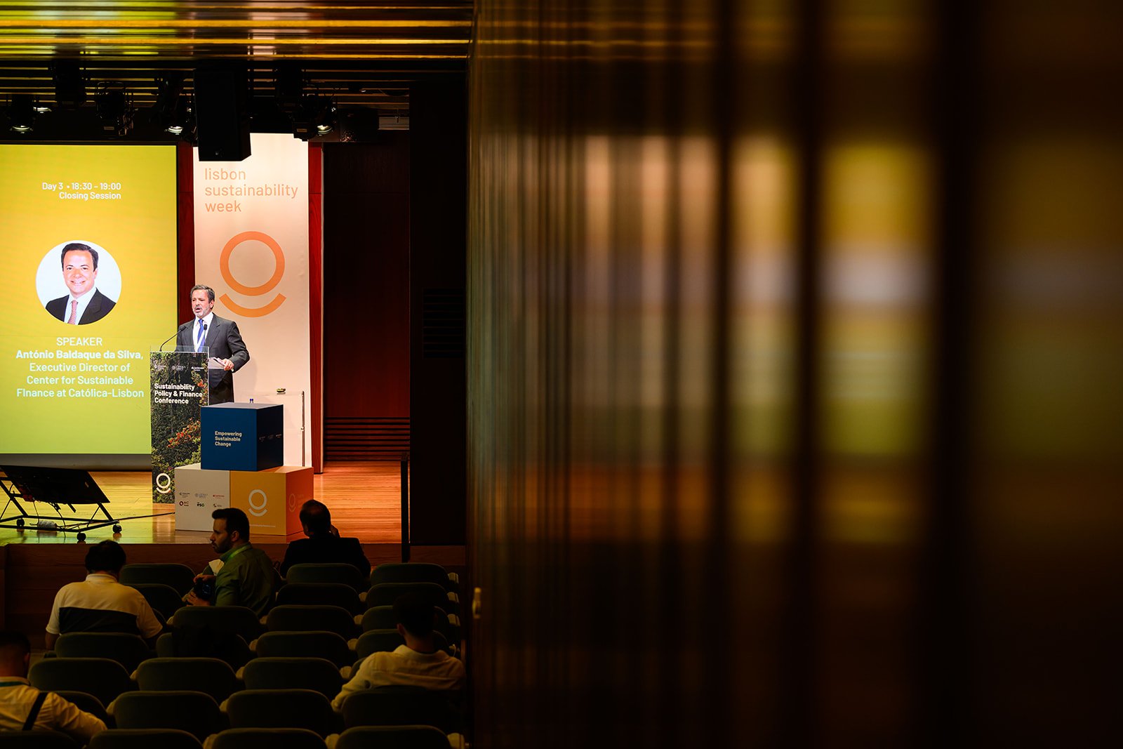 A man is speaking at a podium during a conference called Lisbon Sustainability Week. The screen behind him displays his name as António Baldague da Silva, Executive Director of the Center for Sustainable Finance at Católica-Lisbon.