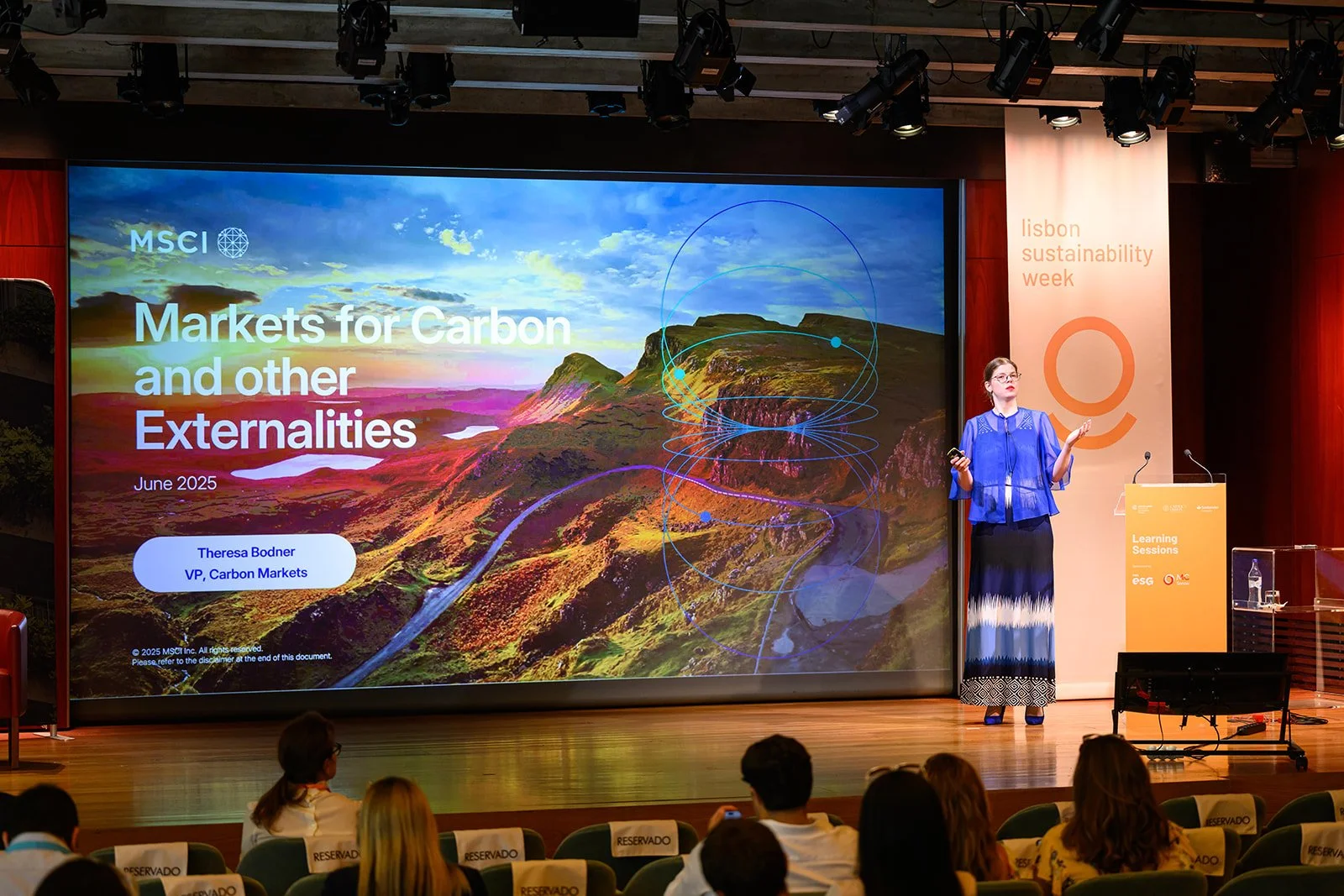 A woman in a blue top and long skirt giving a presentation on stage at a conference with a large screen behind her. The screen displays a landscape with mountains and a road, with the title 'Markets for Carbon and Other Externalities' and the name Th