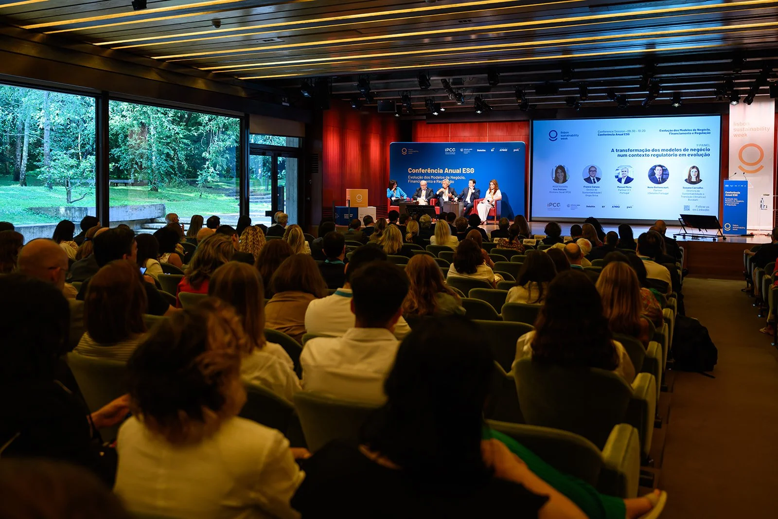 A large group of people attending a conference in a spacious, modern room with large windows showing a lush, green outdoor area. On stage, five speakers are seated, with a blue backdrop and presentation screens displaying conference information.