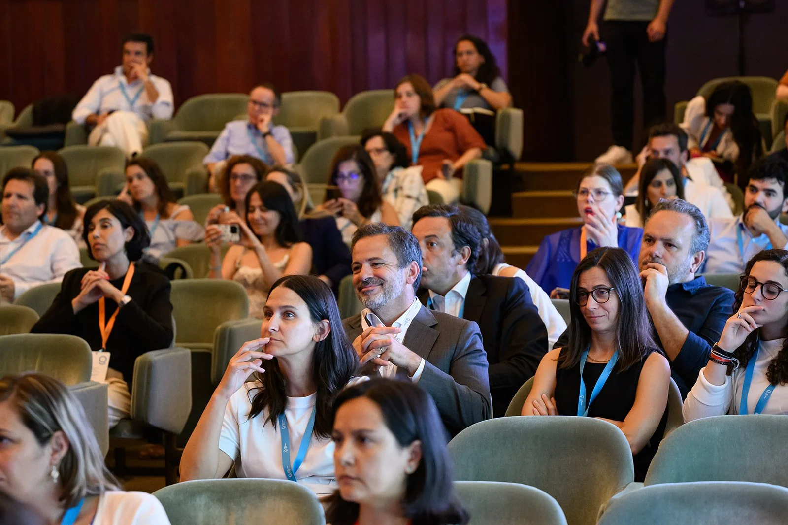 Audience of diverse professionals sitting in conference room, listening attentively, some taking notes or videos, dressed in business casual and formal attire.