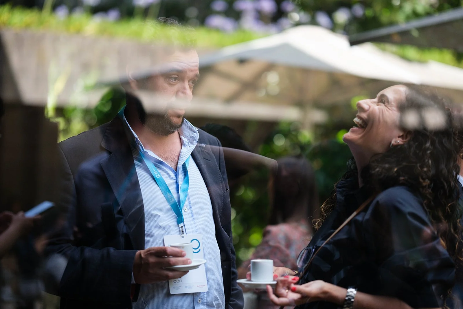 Two people talking outdoors through a glass window, one man and one woman, both holding white coffee cups. The man has a beard, wearing a suit jacket and blue lanyard, the woman has curly hair, laughing and wearing a dark top.