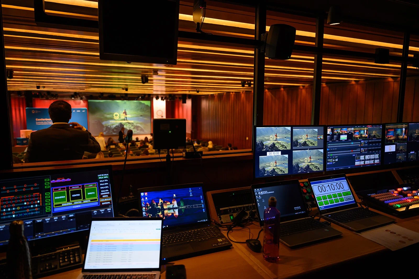Control room with multiple monitors and a view into an auditorium hosting a conference or presentation.