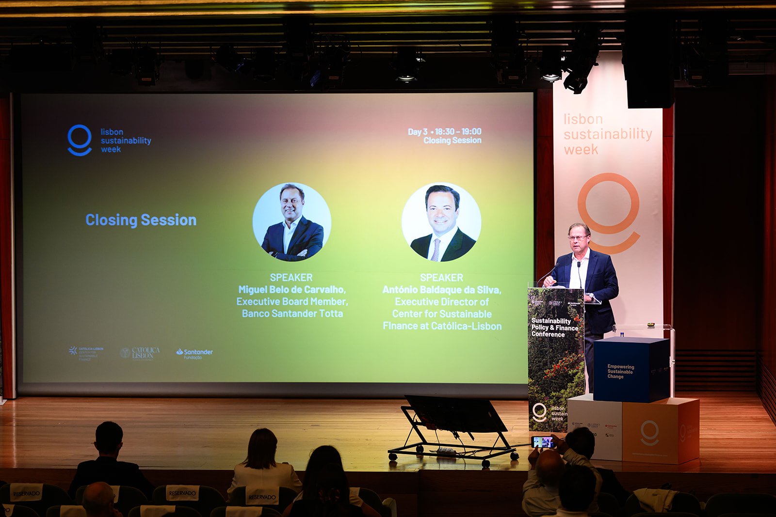 A man in a dark suit at a podium speaking at the Lisbon Sustainability Week, with a large presentation screen behind him displaying details about the closing session featuring two other speakers