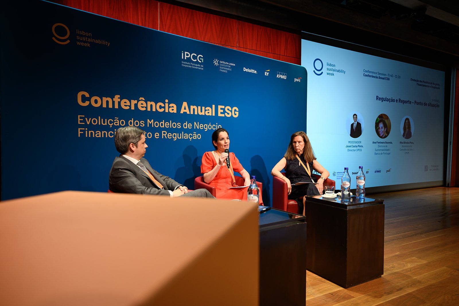Three people speaking on a panel at an ESG conference, with a large blue screen behind them displaying the event's name and details, including logos of sponsors and organizers.
