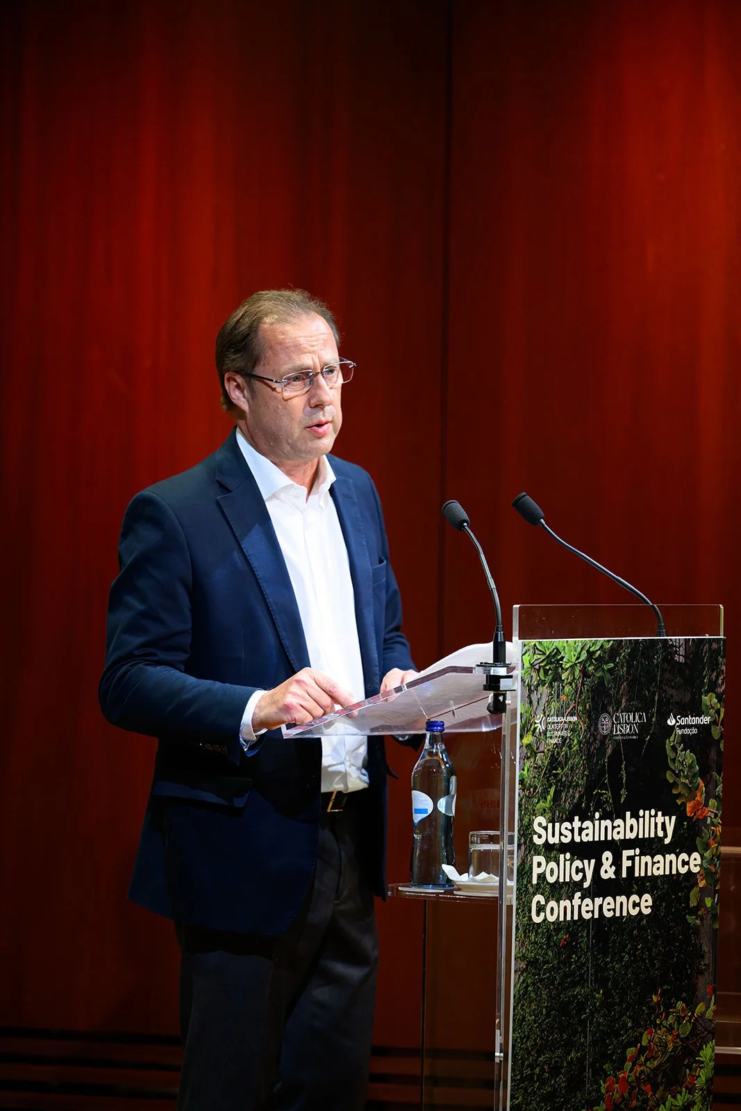 A man in a dark blue blazer and white shirt speaking at a podium during a sustainability policy and finance conference, with a wooden background.
