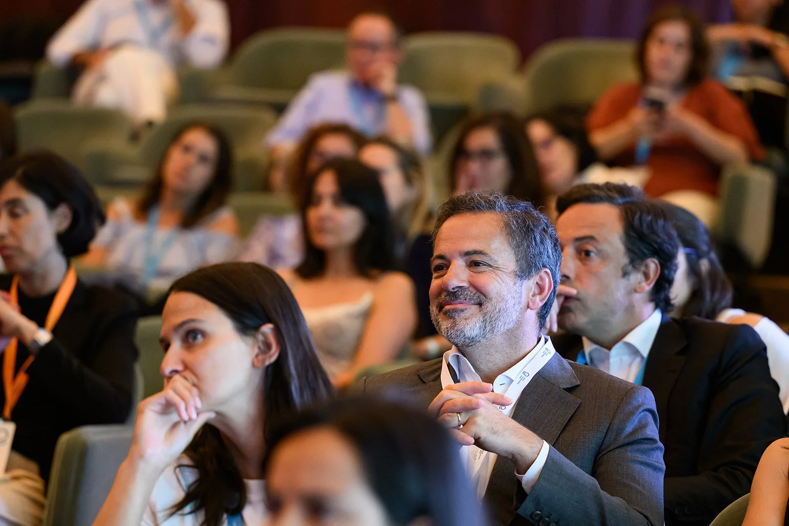 Attendees at a conference or seminar, sitting in green seats, listening and smiling, with some taking notes or using devices.