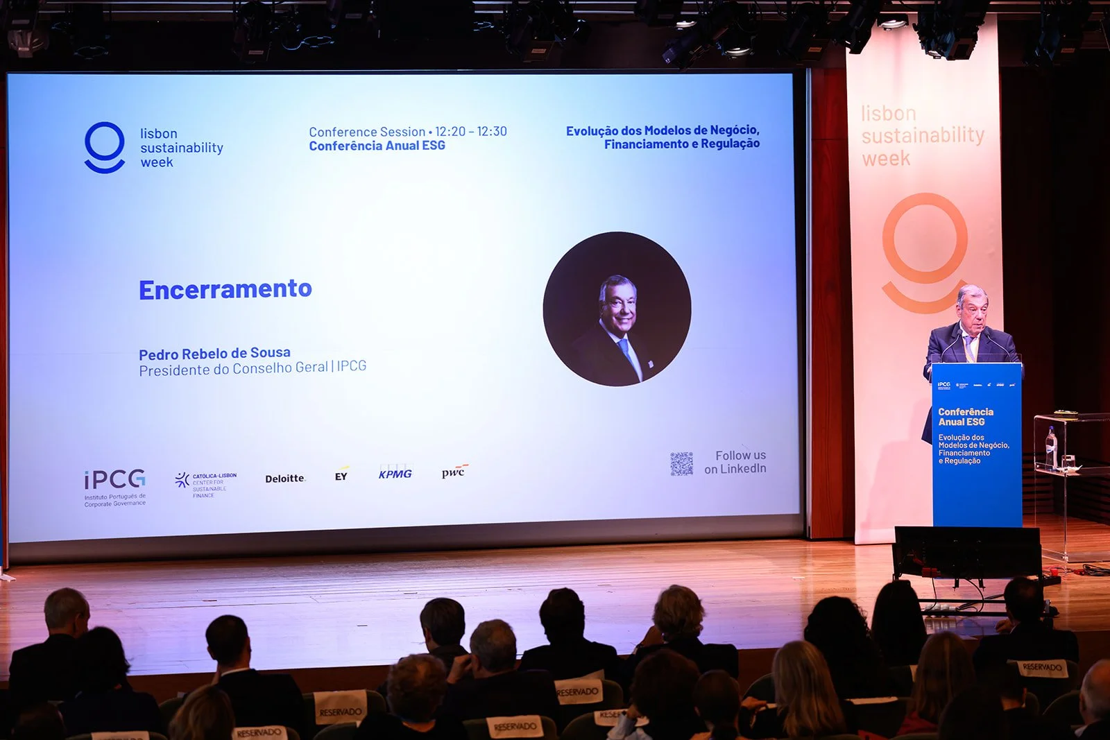 A conference presentation on stage with a large screen displaying the text 'Encerramento' and a photo of Pedro Rebelo de Sousa, with an audience in front and a speaker at a podium to the right.