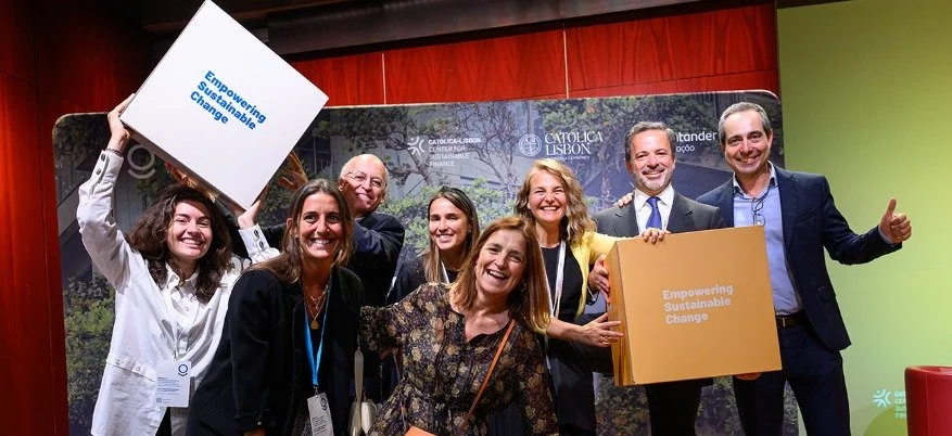 Group of eight diverse smiling people at a sustainability event, holding signs that say 'Empowering Sustainable Change,' with a backdrop featuring 'Catolica Lisbon' and 'Sustainable.'