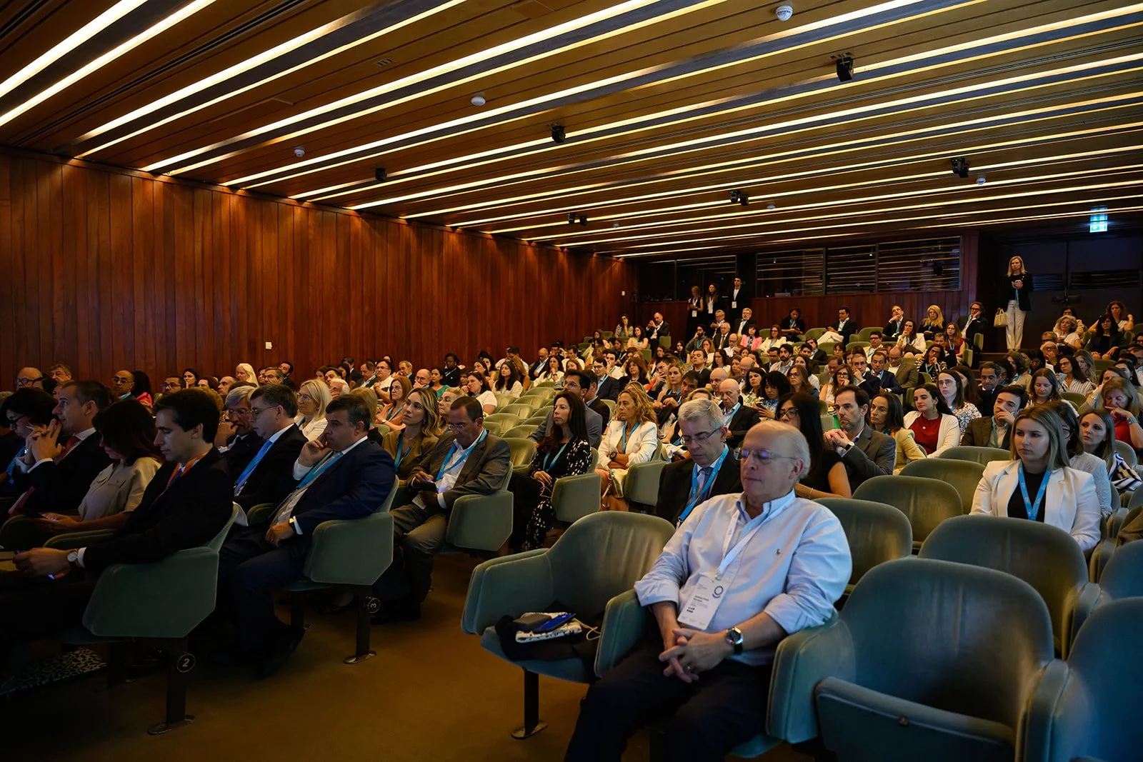 Audience attending a conference or seminar in a large auditorium with wooden panel walls and green chairs.