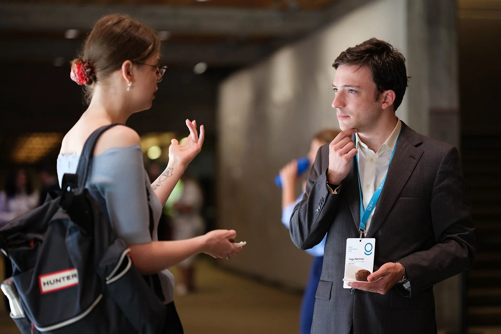 A woman with glasses, a tattoo on her arm, and a backpack is talking with a man in a suit who has a conference badge and a lanyard. He is holding a small plate with food and has a thoughtful expression.