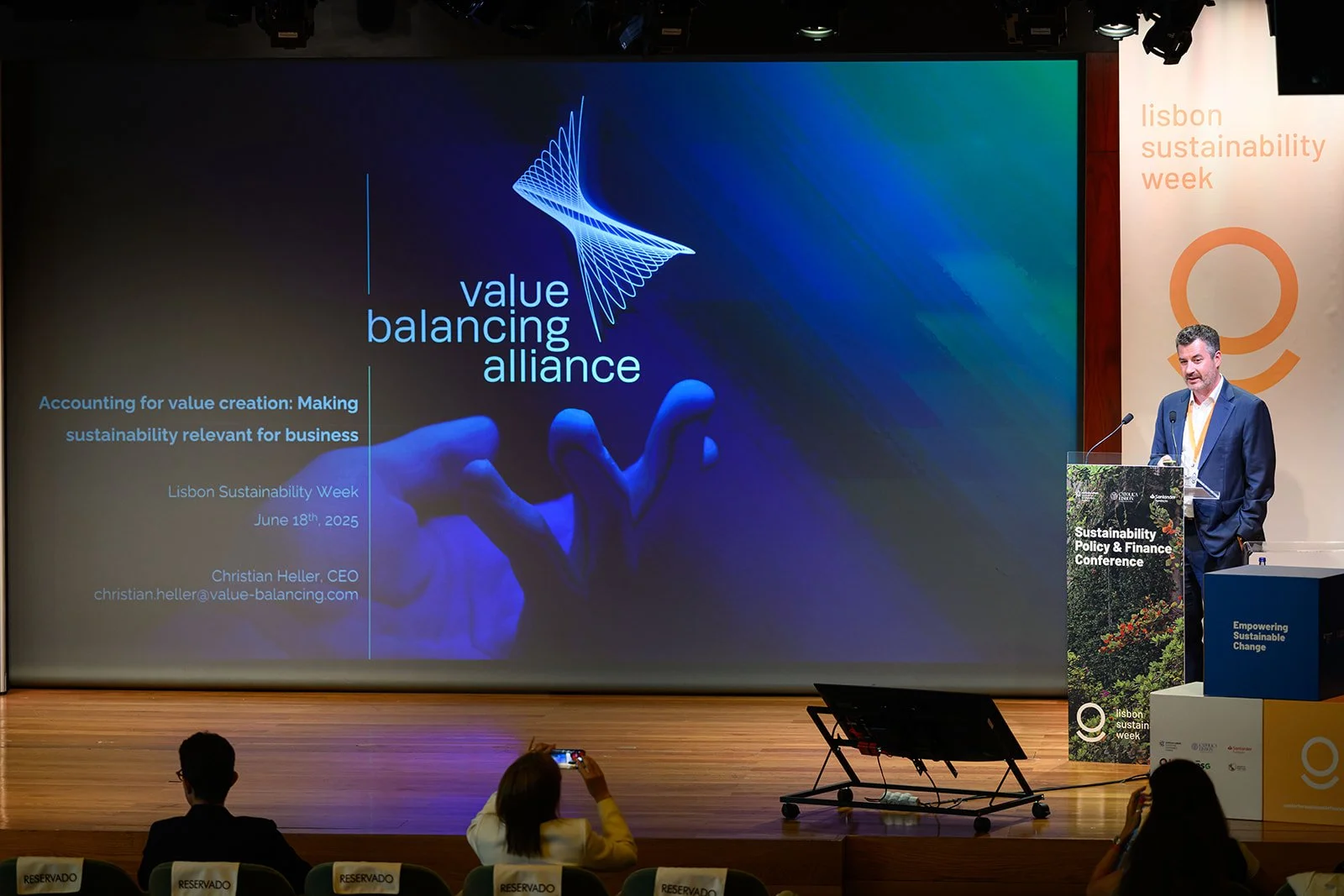 A man in a suit standing at a podium giving a presentation at a conference, with a large screen displaying a slide about value balancing alliance and sustainability week in Lisbon, Portugal.